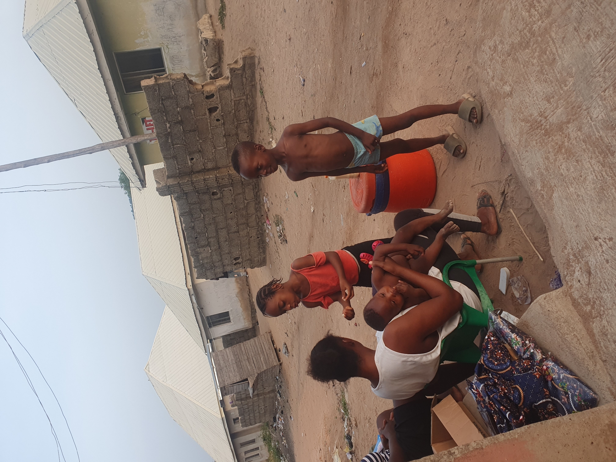 The image shows several people in an outdoor setting in Lokoja, Nigeria. An unpaved area serves as the ground. A young, shirtless boy in light blue shorts stands near an orange and blue plastic bucket. Nearby, a woman in a white tank top and black shorts sits on a green plastic chair. Two other children are seated around her. One is a girl in a red shirt, and the other is a boy with no shirt. Structures of buildings are visible in the background. Trash is scattered around the ground.