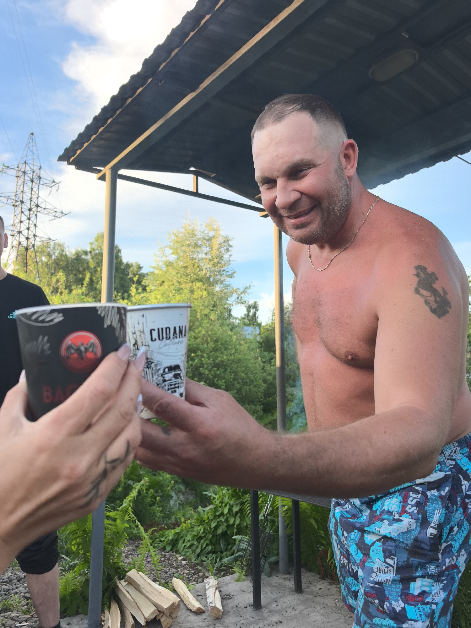 The image captures an outdoor gathering in Noviy, Russia, featuring a smiling man toasting with another individual. The man, shirtless with a short haircut, light beard, and a gold chain, wears blue and white patterned swim shorts with text like "SPORT," "JOSS," "SWIMMING," "ACTIVE," and "WATER." A dark tattoo resembling a figure is visible on his left shoulder. He is holding a white disposable cup adorned with the text "CUBANA" and a cityscape illustration. His cup is clinking with a black disposable cup held by another person's hands, which features the "BACARDI" logo and the text "BA." Part of another person, wearing a black t-shirt with a partial "adla" (likely Adidas) logo, is visible on the far left.
The setting is outdoors under a dark corrugated canopy or shelter, with a metal support pillar visible. Logs of firewood are stacked on the ground nearby, and a metal grill grate hangs to the side, suggesting a barbecue or fire pit activity. The background shows lush green foliage and trees under a bright blue sky with some clouds. A large power line tower is visible in the distance. The scene is well-lit by daylight, indicating clear weather. The man's broad smile conveys a relaxed and happy mood during this informal outdoor event.