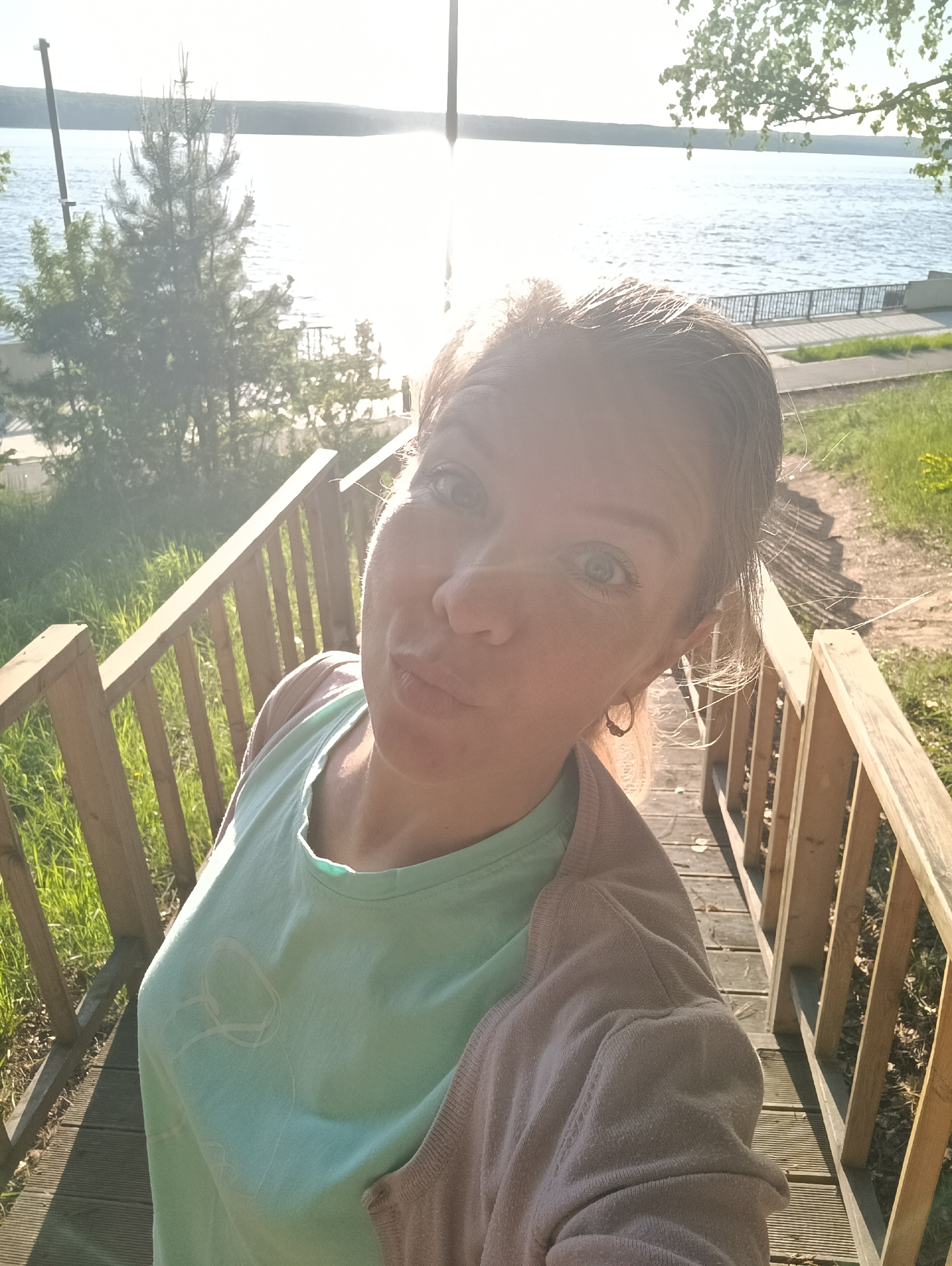 A selfie was taken on a wooden deck next to the lake. A woman stands on wooden steps with the wooden railing around her. She has blue eyes and reddish-blond hair pulled back in a ponytail. She is wearing a light blue t-shirt with a light pink or beige cardigan. The area around her is green with foliage and grass. A body of water appears to be a lake. The location appears to be a recreation area or park in the city of Chaikovsky, Russia.