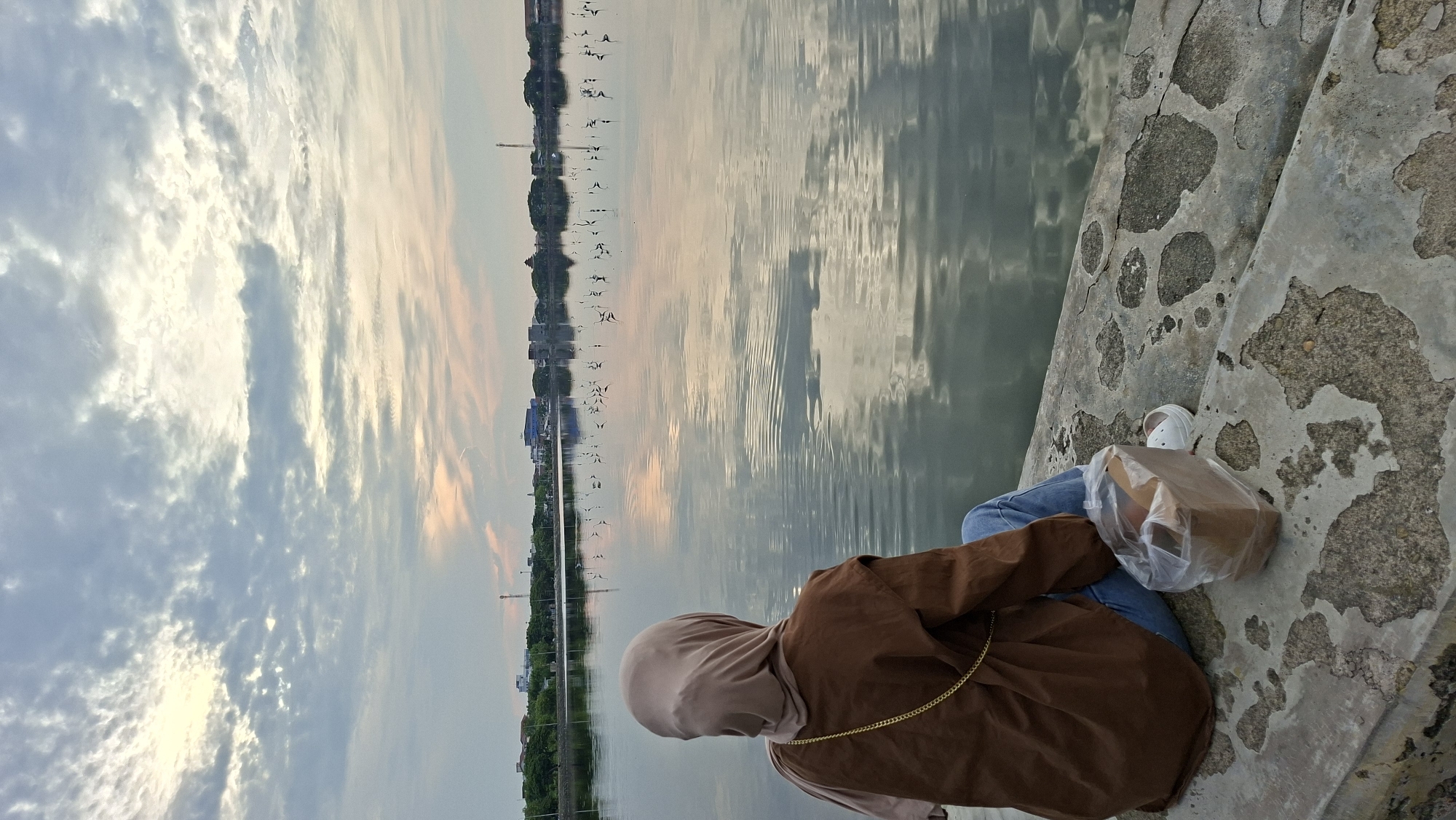A person wearing a hijab and a brown jacket sits on a concrete ledge overlooking a body of water. The person is seen from behind, and their legs are crossed, with blue jeans and white shoes visible. Next to the person is a plastic bag containing a brown paper box. The water reflects the sky, which is filled with clouds, and a distant cityscape with buildings and trees can be seen across the water. The concrete ledge has a textured, worn appearance. This scene is likely at the Tugu Km 100, a landmark and recreational area in Lhokseumawe, Indonesia, known for its waterside views.