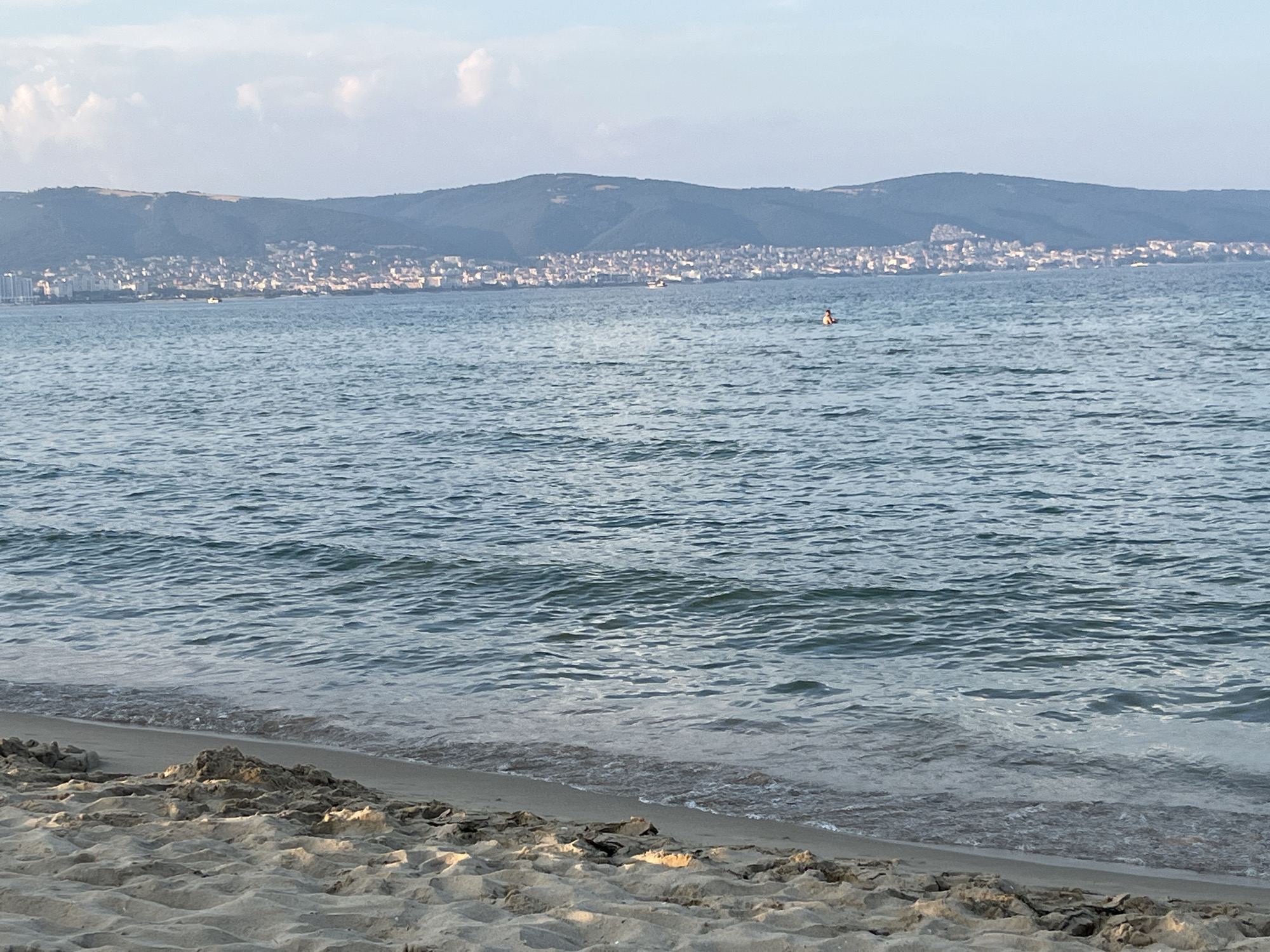 The image depicts a coastal scene in Nesebar, Bulgaria, featuring a sandy beach in the foreground. Gentle waves lap the shore, and the sea stretches out towards a distant, hazy coastline. A small figure is visible in the water, appearing to be swimming or wading. The background showcases a sprawling town nestled against a backdrop of rolling hills, indicative of Nesebar's unique geography. The sky is partly cloudy, suggesting fair weather. The overall atmosphere is serene and suggestive of a summer day at the beach.