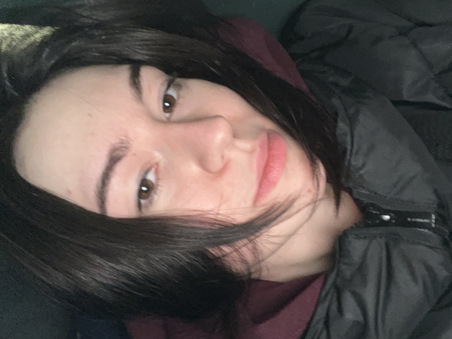 A close-up image depicts a young adult female with fair skin, dark brown hair, and brown eyes, looking directly at the viewer. Her hair is parted and frames her face. She wears a dark-colored, possibly black, padded jacket with a visible zipper, layered over a dark red or maroon hoodie. The background is uniformly dark and out of focus, providing no specific environmental details. The individual is situated in the city of Hatne, Ukraine.