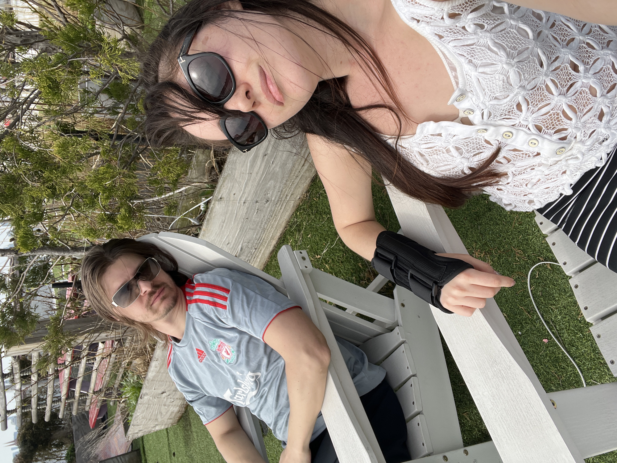 The image shows a fair-skinned woman with long brown hair and sunglasses lying on a wooden bench or structure. She is wearing a white, floral-patterned top. Next to her, a fair-skinned man with long brown hair and sunglasses sits in a white Adirondack chair. He is wearing a grey Liverpool football shirt with red accents. The woman has a black brace or splint on her left wrist or hand. The scene is outdoors, with green grass and a glimpse of a body of water and buildings in the background. The location is not identifiable in the image as being within New York City.
