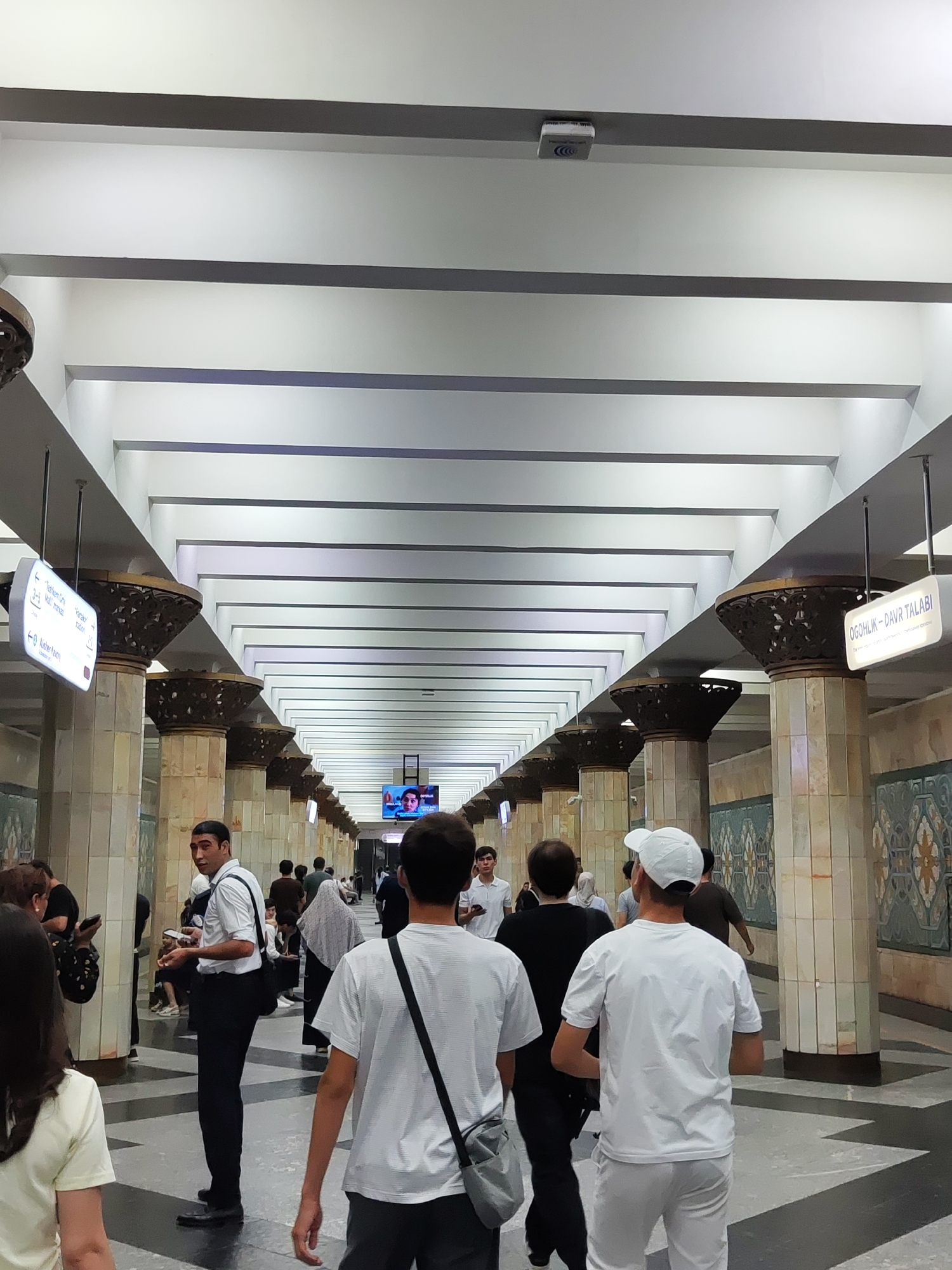 The image shows the interior of a subway station in Tashkent, Uzbekistan. The station features a long, vaulted ceiling with parallel light fixtures. Large, ornate pillars line the sides of the platform, and decorative tiling adorns the walls. Several people are walking along the platform, some in groups and others individually. Signs in a foreign language are visible, indicating station names and directions. In the foreground, two individuals are walking away from the camera, and another person is looking back over their shoulder. The overall impression is of a busy public transportation hub.
