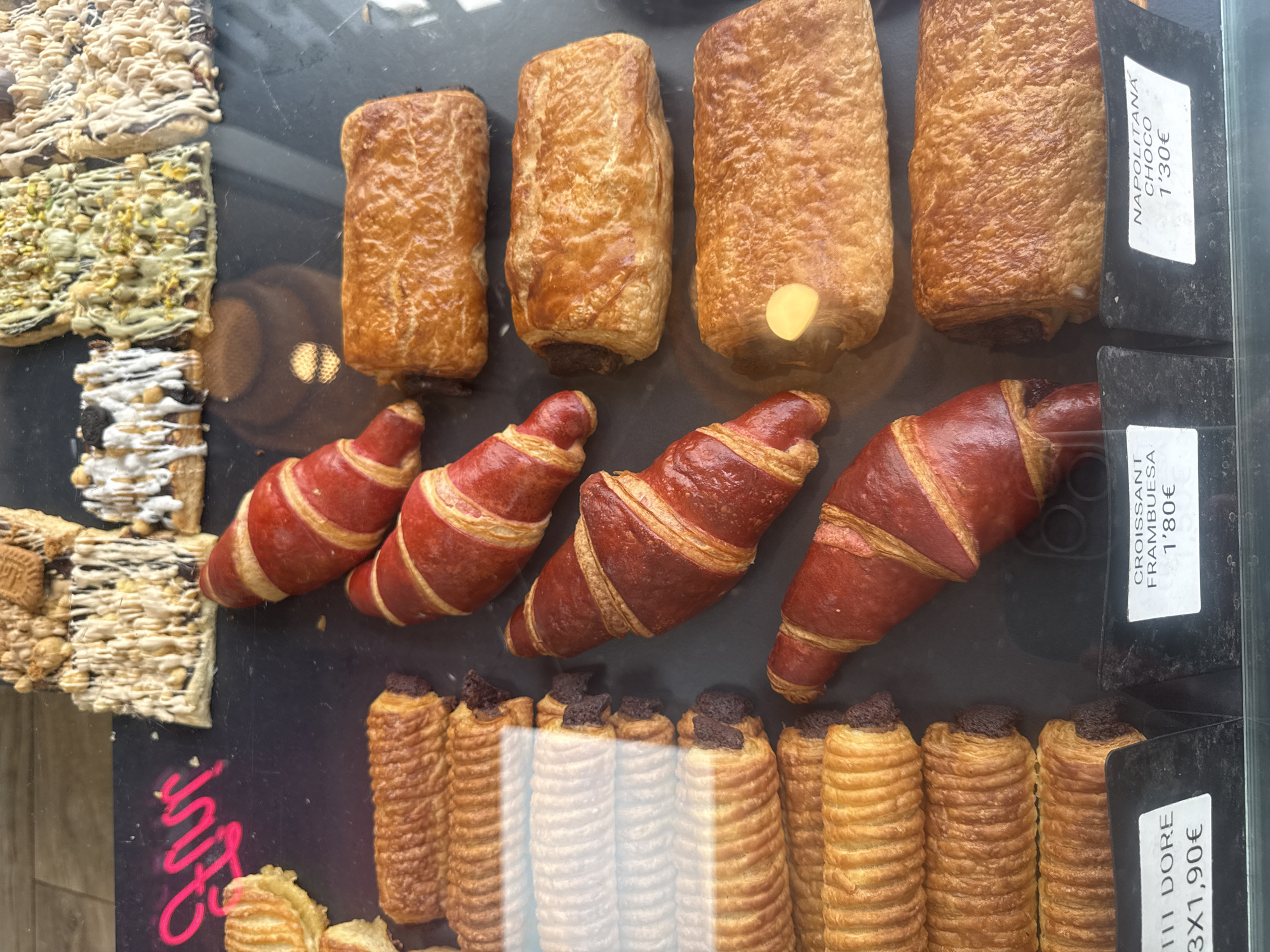The image displays a variety of baked goods neatly arranged in rows within a glass display case, likely in a bakery or patisserie setting. The items are well-lit, highlighting their golden-brown and reddish hues, and appear fresh and appetizing.
In the top row, there are four rectangular, golden-brown pastries with a flaky, puffed appearance, possibly resembling pain au chocolat. Below these, a middle row features four distinctive croissants. These croissants have a reddish-brown color with lighter golden-brown stripes, suggesting a specific flavor or glaze, such as raspberry.
The bottom row showcases several cylindrical pastries with a ridged texture, each topped with a dark chocolate chunk or spread. These could be an elongated version of pain au chocolat or similar sweet rolls. On the far left, a selection of flat, rectangular bar-shaped pastries or cookies is visible. These are adorned with various toppings, including white icing with nuts, green pistachio pieces, and chocolate drizzles.
Several vertical white labels with black text provide information about the products and their prices. At the top right, a label reads "NAPOLITANA CHOCO 1'30€", indicating a chocolate Neapolitan pastry priced at 1.30 Euros. Directly below it, another label states "CROISSANT FRAMBUESA 1'80€", identifying the red croissants as raspberry-flavored and costing 1.80 Euros. At the bottom right, a third label reads "III DORE 3X1,90€", possibly referring to the ridged pastries as "Dore" and priced at three for 1.90 Euros. A partially visible, bright pink neon-like drawing or text can also be seen on the dark surface in the bottom left corner.