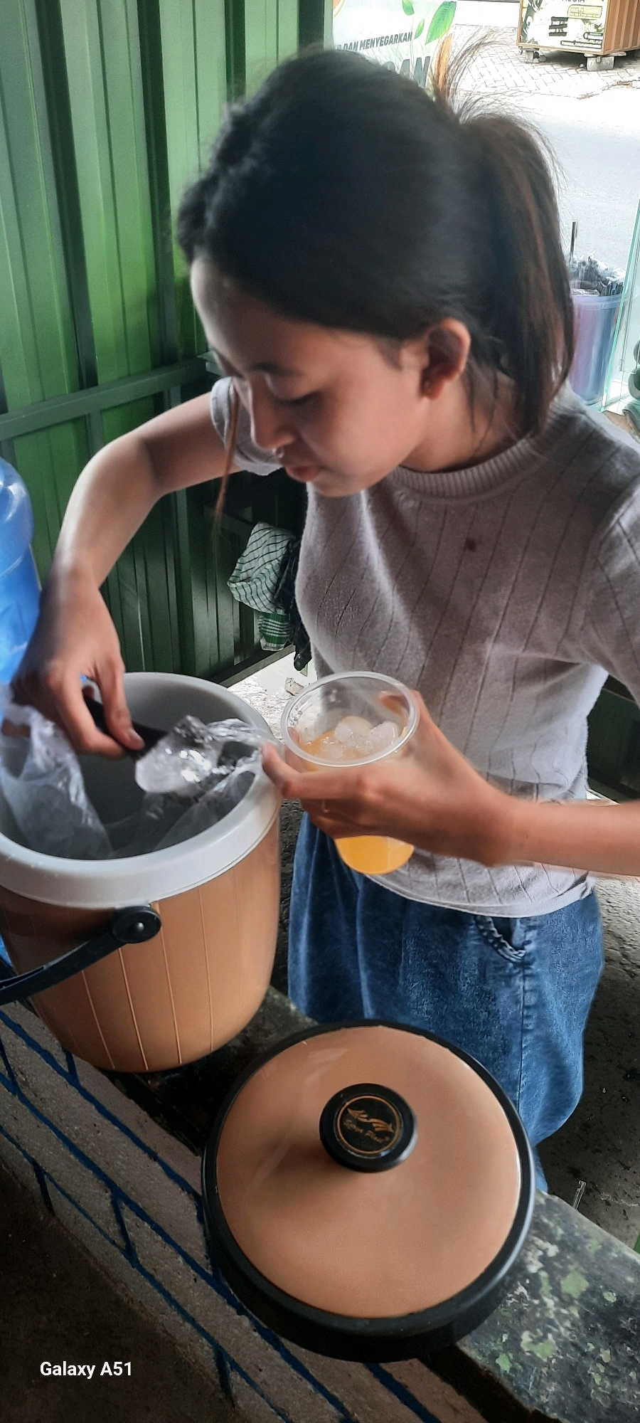 A young woman with dark hair pulled back in a ponytail, wearing a grey ribbed sweater and a blue denim skirt, is standing in what appears to be an outdoor food stall or market area in Selopuro, Indonesia. She is in the process of preparing a drink. In her left hand, she holds a clear plastic cup filled with ice and an orange beverage, likely juice or tea. Her right hand is reaching into a large, light brown plastic cooler with a white rim, holding a clear plastic bag containing more ice and possibly scoops. The cooler is on a weathered wooden counter. Behind her, there is a green corrugated metal wall and a glimpse of a white sign with text that includes "DAN MENYEGAR", and a blue water cooler jug. In the foreground, a round, copper-colored lid with a black knob featuring a logo is placed on the counter. The overall impression is a casual, everyday scene of drink preparation.