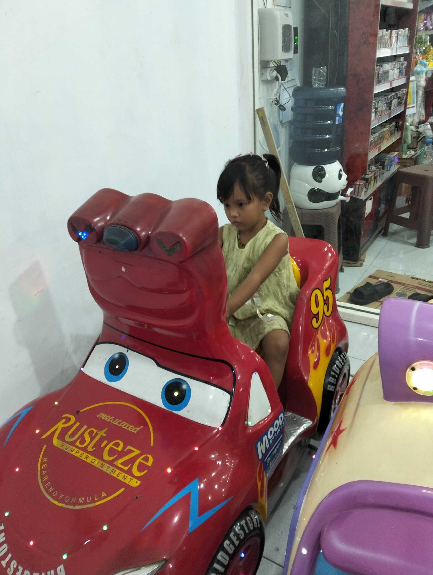 A young Indonesian girl with dark hair tied in a ponytail sits in a red toy car resembling Lightning McQueen from the movie "Cars". She wears a light green, sleeveless dress. The toy car is red with blue eyes, yellow flames, and the number "95" on its side. The "Rust-eze" logo, which is the brand of bumper ointment from the movie, is visible on the car's hood. The car has LED lights that are illuminated.
The girl is seated in what appears to be an indoor establishment, possibly a game arcade or a shopping mall in Kedungrejo, Indonesia. In the background, there is a water dispenser with a panda-themed base, a shelf stocked with various items, and a brown stool. The flooring is tiled. The environment suggests a public space designed for entertainment or commerce.