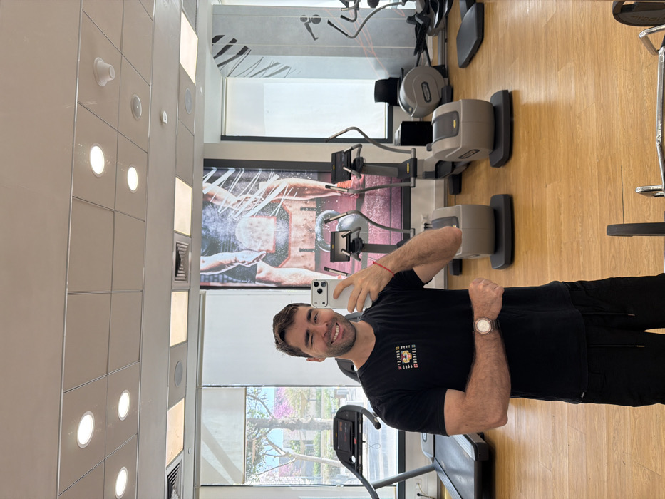 A male individual is depicted in a mirror selfie taken within a modern fitness center in Tel-Aviv, Israel. The subject, a light-skinned man with short brown hair and a beard, is smiling broadly and winking with his left eye. He holds a light-colored smartphone in his right hand, positioned to capture the image. His left arm is bent, with his hand clenched into a fist at waist height. He wears a black t-shirt with a colorful graphic design on the chest, black shorts or sweatpants, a light-colored wristwatch on his left wrist, and a red string bracelet on his left wrist.
The environment features a light wooden floor and a white paneled ceiling with multiple recessed circular lights. Various gym equipment is visible in the background, including a black treadmill on the far left and multiple light-grey elliptical machines (Technogym branded) arranged in the mid-ground. A black fitness mat rests on the floor to the right. A large wall banner behind the equipment displays a partial image of a person exercising with a kettlebell, with white chalk dust particles depicted in the air. Through large windows on the left, an outdoor scene is visible, featuring green foliage and trees with pink blossoms, suggesting an urban or park setting. A barbell or weight rack is partially visible in the foreground at the bottom of the image.