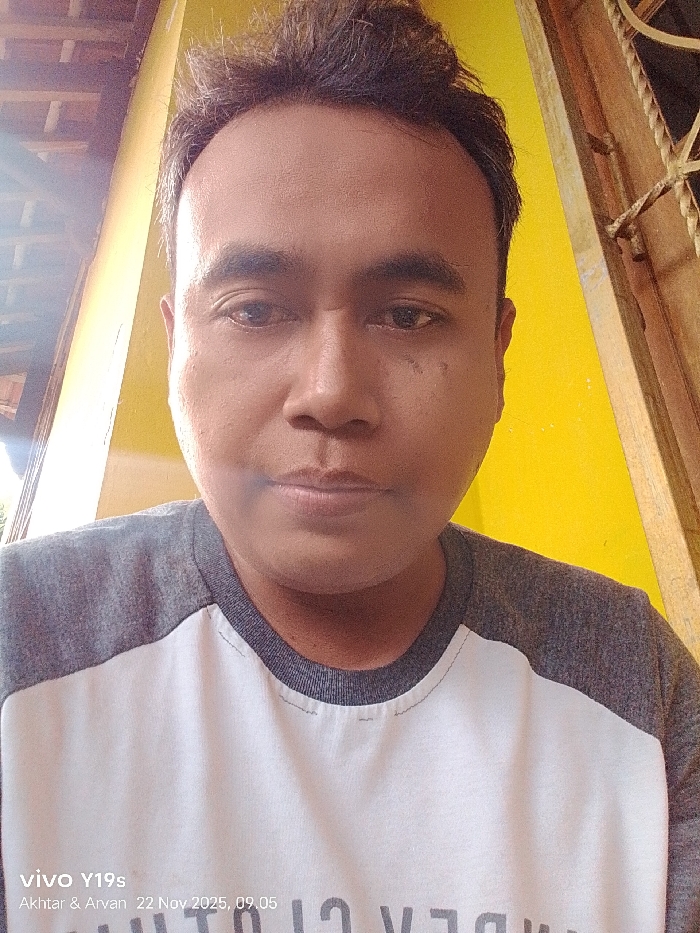 A person with short, dark hair and tan skin is centered in the frame, facing forward. They have a neutral expression. They are wearing a grey raglan-sleeved shirt with a white front panel. The background includes a bright yellow wall and a window with decorative metal bars. The lower left corner of the image displays "vivo Y19s Akhtar & Arvan 22 Nov 2025, 09.05". The image appears to be a selfie taken indoors.