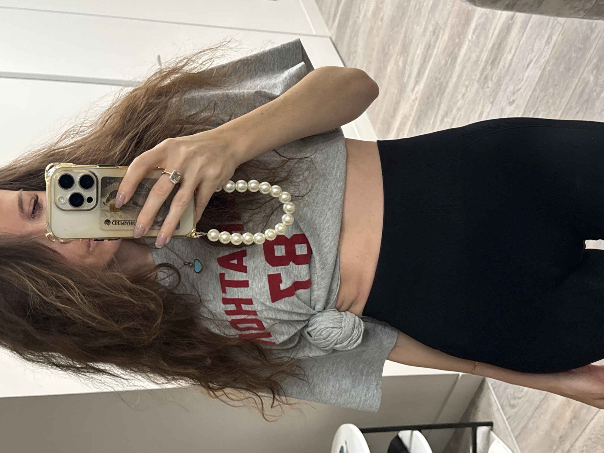 A woman is taking a selfie in a room with white walls and light wood-patterned flooring. She is wearing black leggings and a gray t-shirt with red lettering, knotted at her waist. She holds a gold-colored smartphone with a pearl beaded strap. A piece of white furniture is visible in the lower right corner. The city of Khimki, Russia is not discernible from this image.