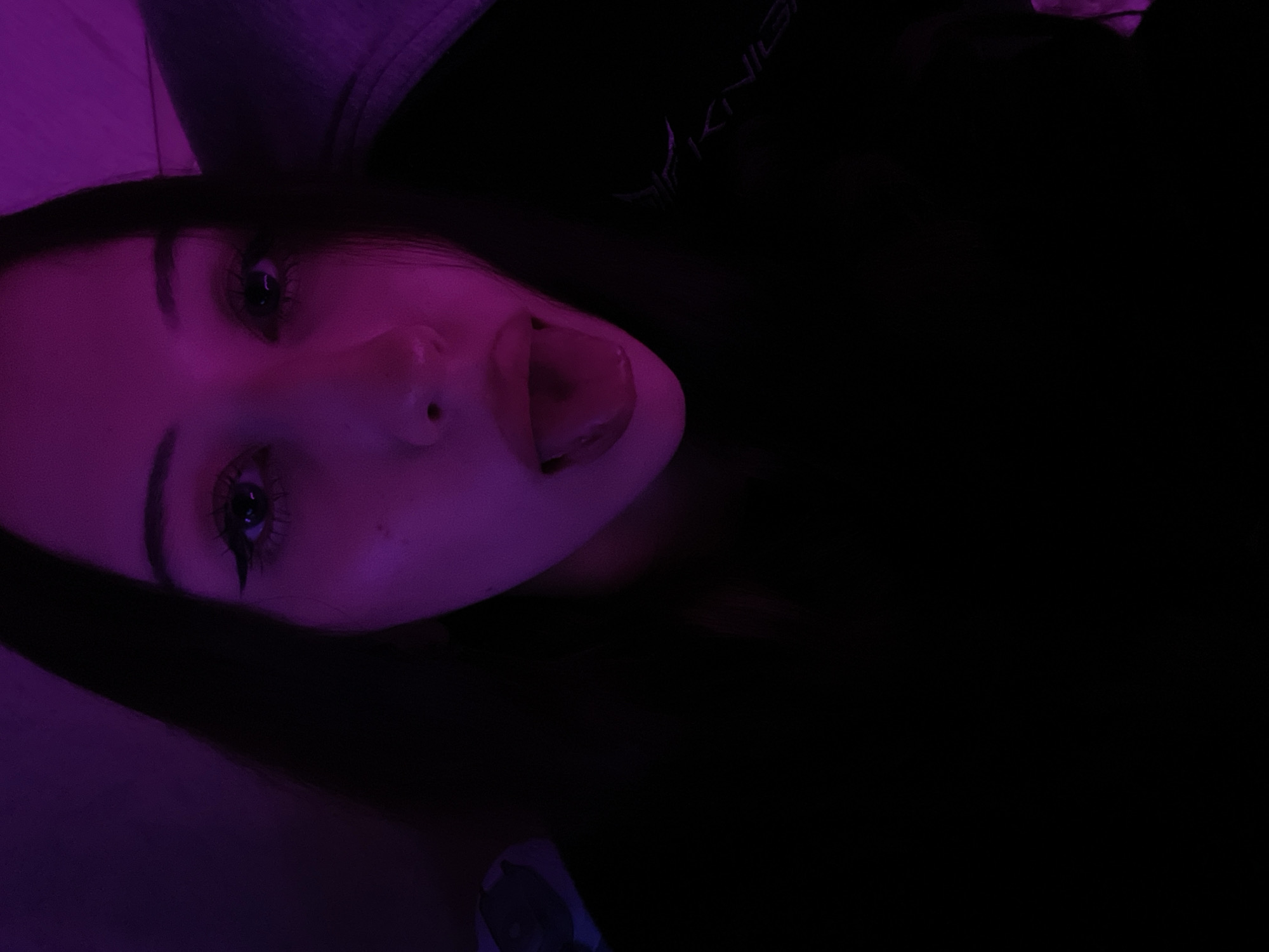 The image displays a headshot of a young individual, likely female, illuminated predominantly by a strong magenta-purple light source from the front-right. The subject is centered in the frame, directly facing the camera, with their mouth open and tongue extended.
The individual has long, dark hair, which is partially visible around the face. They are wearing dark (likely black) clothing. On the lower right side of the frame, a partial, stylized logo or text in a lighter color is visible on the garment, appearing to spell "KNIGHT" or a similar word, with a prominent "K" or "R".
Facial features are clearly discernible despite the color cast. Both eyes have prominent winged eyeliner. A small dark mole or freckle is visible on the subject's right cheek (viewer's left), below the eye. The background is dark and indistinct, suggesting an indoor setting, with faint, diffuse purple light visible on what appears to be a surface in the upper right portion of the frame.
The image provides no discernible visual information to indicate a specific geographic location such as Votkinsk, Russia. The setting appears to be a generic indoor environment.