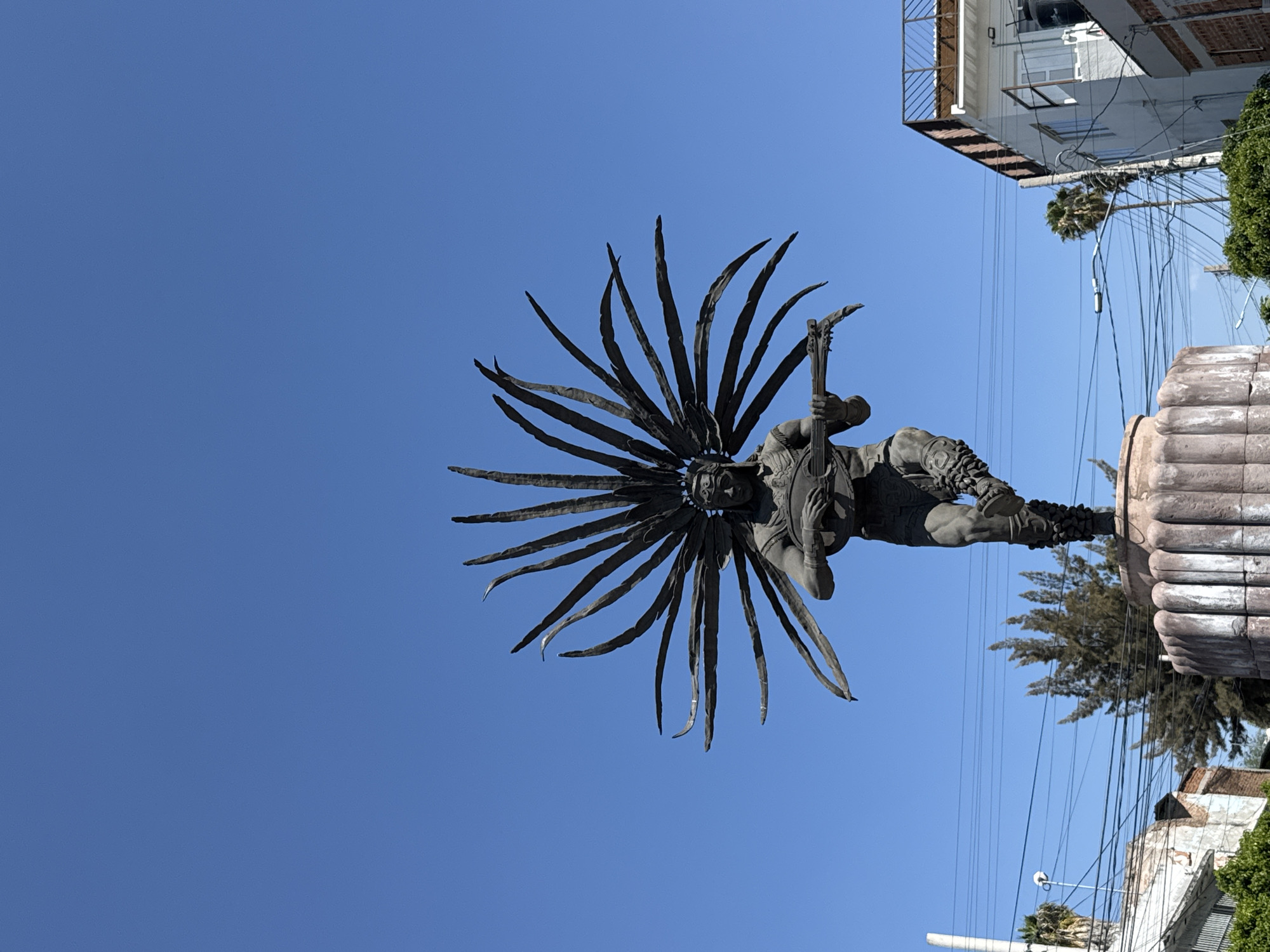 A low-angle, rotated view captures a statue of an indigenous man with a headdress, holding a stringed instrument, likely a guitar, in Querétaro, Mexico. The statue stands atop a textured, cylindrical base, set against a clear blue sky. Power lines intersect the view and blend with the branches of a tree. Parts of a building with a balustrade can be seen in the top right corner.
