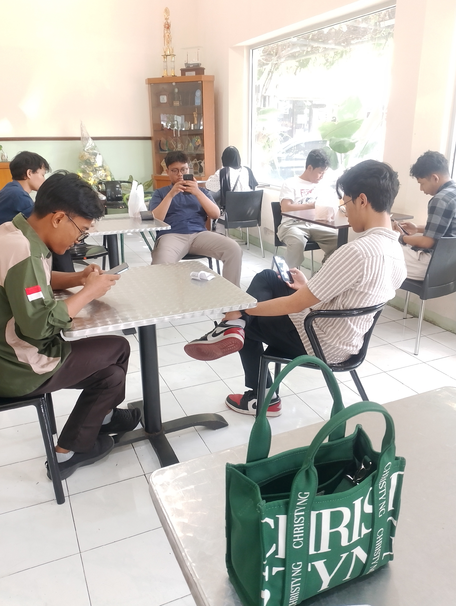 The image depicts a group of young men sitting at tables in a bright, indoor space with white tiled floors and light-colored walls. The room appears to be a cafe or dining area, with natural light streaming in from a large window at the back, revealing greenery outside. In the background, there is a wooden display cabinet filled with trophies and awards, suggesting the location might be associated with an educational institution or club.
Several individuals are seated, each engaged with their mobile phones. One person in the foreground, wearing a green and tan jacket with an Indonesian flag patch, is looking down at his phone while seated at a metal-topped table. To his right, another person in a striped shirt is also looking at his phone, seated in a black chair. Further back, other young men are visible, also engrossed in their devices. A green tote bag with "CHRISTYNG" written on it is placed on a table in the foreground. The overall atmosphere is one of casual use of technology in a communal setting. Given the context of trophies and the presence of young people, it is plausible this scene is located within a university or college campus in Yogyakarta, Indonesia.