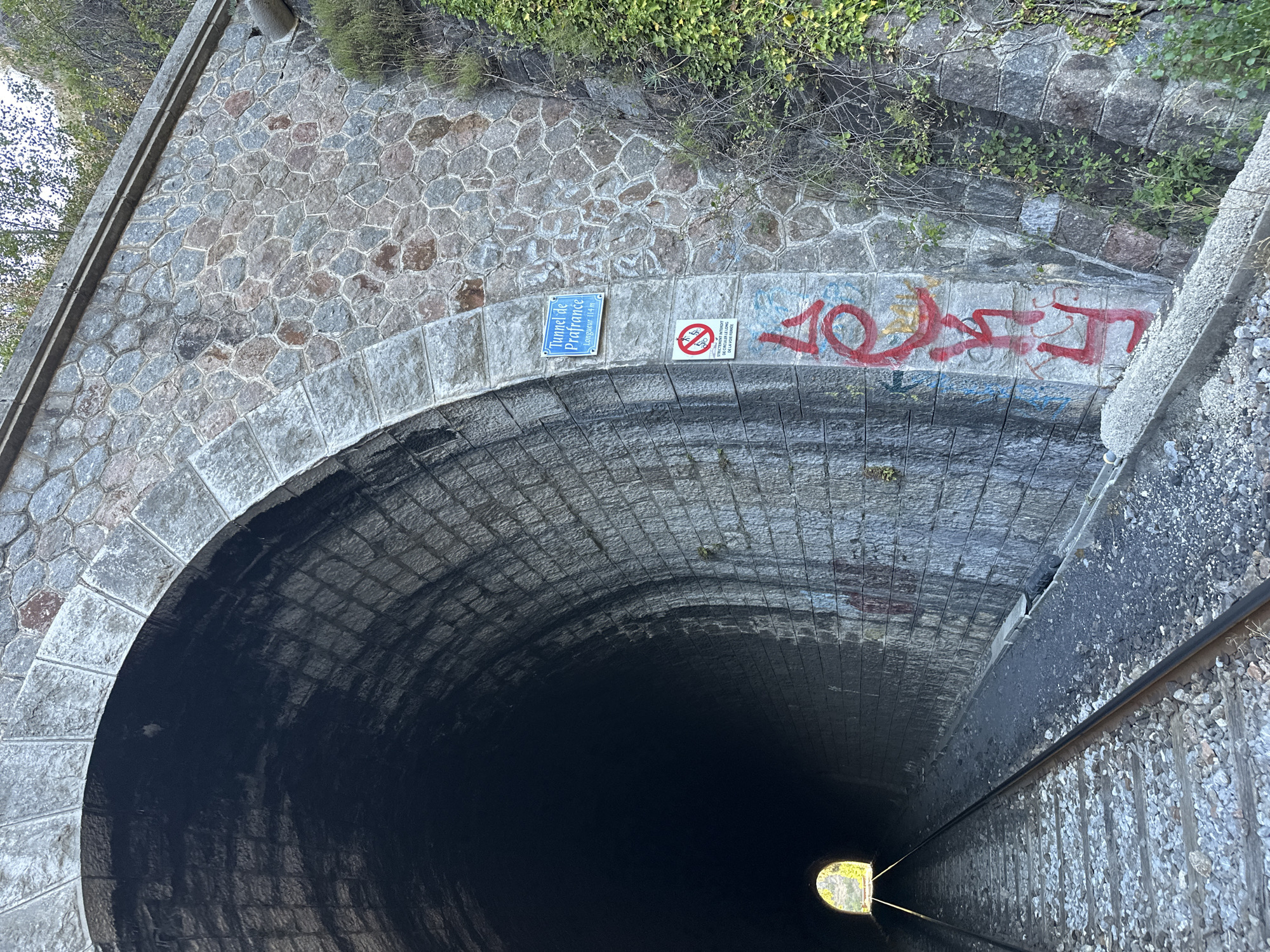 The image shows the entrance to a stone tunnel, identified by a blue sign as "Tunnel de Prafrance, Longueur 114m". The tunnel is part of a railway line, with tracks visible in the foreground entering the dark interior. The tunnel walls are constructed of rough-hewn stone blocks, with some sections showing signs of age and weathering. Graffiti is present on the exterior wall of the tunnel, including a red tag that appears to read "LOU". To the right of the entrance, ivy and other vegetation grow up the stone embankment. A circular sign with a red prohibition symbol indicates that bicycles are not allowed. The scene is outdoors, with trees visible at the top of the tunnel's entrance. The location is identified as Générargues, France.