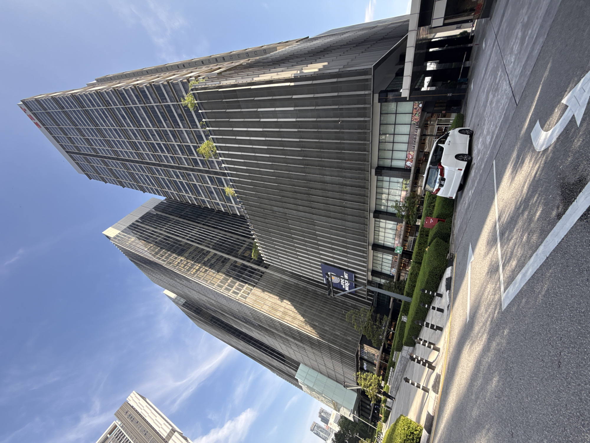 The photograph depicts two tall, modern skyscrapers against a clear blue sky with some wispy clouds. The buildings feature a repetitive pattern of dark horizontal lines or panels and glass. A white van is parked beside the building, near a sidewalk with meticulously trimmed green hedges. A road marking with an arrow is painted on the grey asphalt. The perspective suggests the photograph was taken from a low angle, emphasizing the buildings' height. The presence of a sign in the background suggests the location is in Kuala Lumpur, Malaysia.
