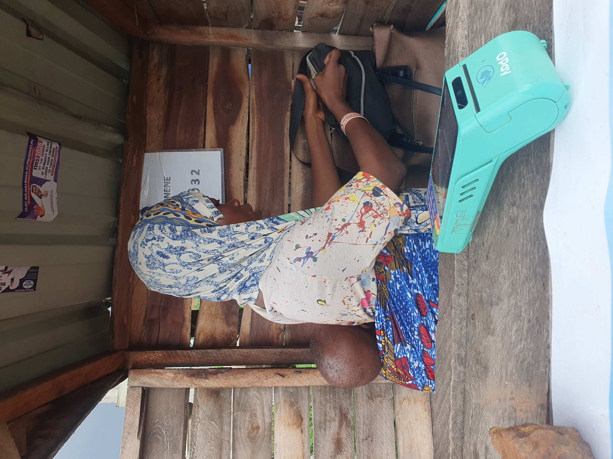 A medium shot captures a woman wearing a blue and white patterned headscarf and a paint-splattered shirt, seated inside a wooden stall in Lokoja, Nigeria. She is facing to the right, with her left arm resting on a black bag and her right hand reaching into it. Next to her, a baby is visible, head resting on her shoulder. A turquoise Opay card reader is positioned on a wooden surface in the foreground. Behind the woman, a sign with the name "ENENE" and numbers "32" is attached to the wooden wall. The stall has a corrugated metal roof and wooden plank walls.
