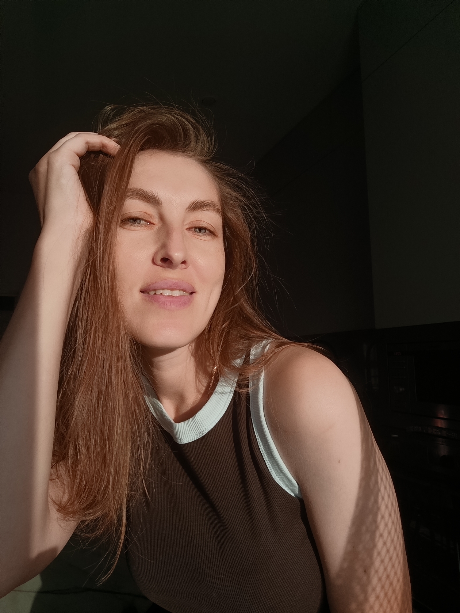 A woman with long, auburn hair is captured in a close-up, looking towards the camera with a gentle smile. Her hand is raised, resting on her head and partially obscuring her hair. She wears a brown tank top with a light blue trim. The scene is illuminated by warm, low-angled sunlight, casting shadows across her face and arm, suggesting it is either early morning or late afternoon. The background is dark and out of focus, with a hint of what appears to be a kitchen appliance on the right side. The overall mood is relaxed and natural, possibly taken in a home environment. There is no visible text in the image.