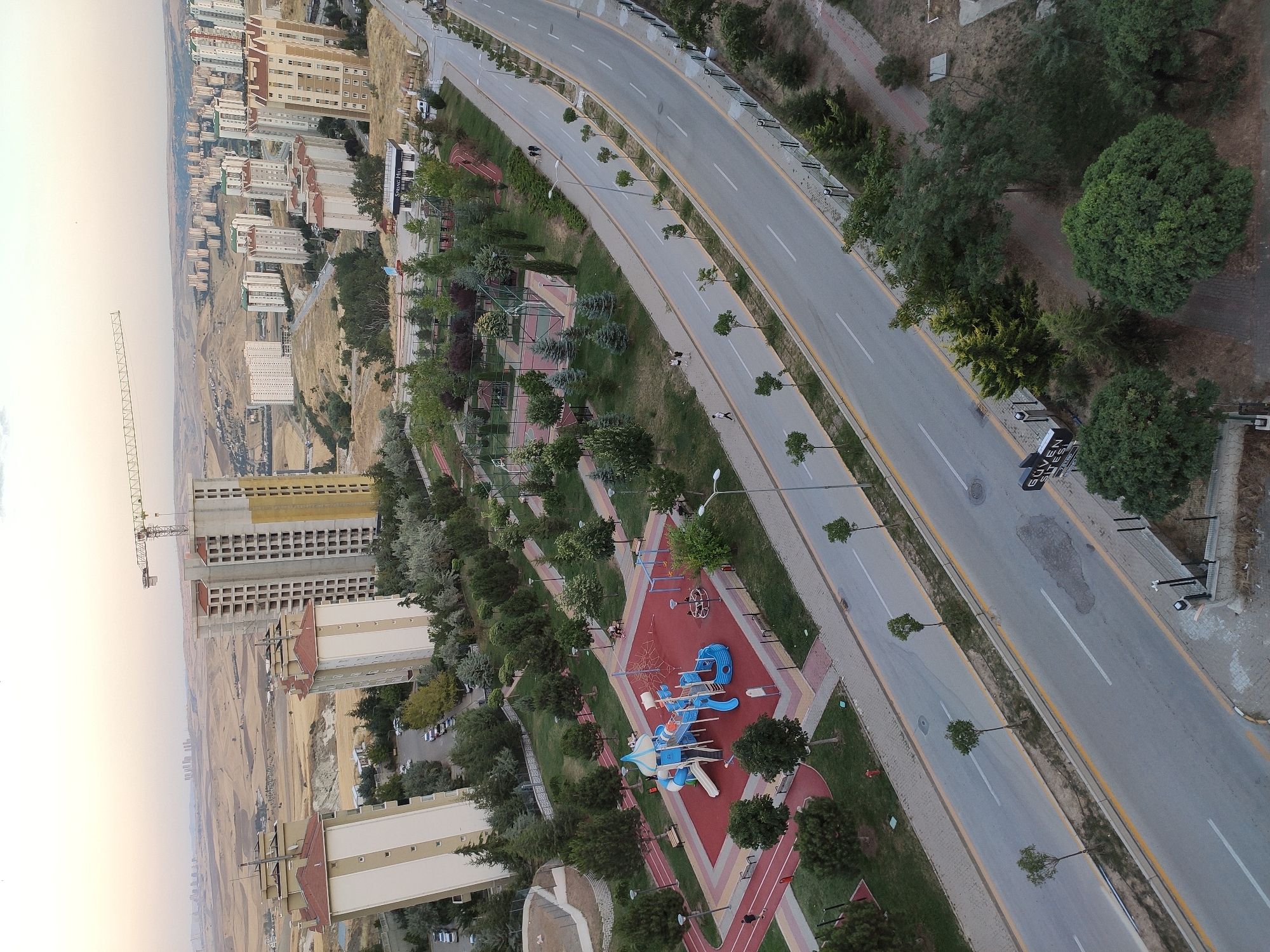 An aerial view captures a developing urban landscape in Etimesgut, Turkey, at dusk. The scene is dominated by several multi-story apartment buildings, some completed and occupied, others under construction with cranes visible on their upper floors. A wide, paved road with yellow lane markings runs through the center of the image, bordered by sidewalks and trees. On one side of the road, a park features a vibrant red playground with blue and white climbing structures, slides, and swings, surrounded by green grass and trees. Further into the park, basketball courts and covered seating areas are visible. People are observed walking along the sidewalks and in the park. The background reveals a sprawling city with numerous residential buildings stretching towards a hazy horizon, interspersed with arid, hilly terrain. The sky exhibits a soft gradient of light colors, indicative of the setting sun.
