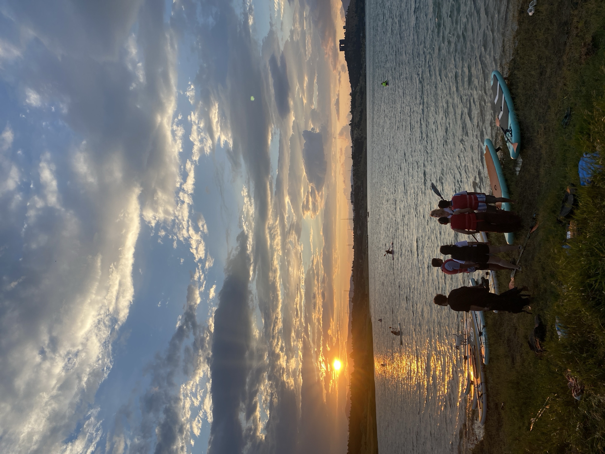 This is a vertical, eye-level, full shot of people with paddleboards at sunset near Tbilisi, Georgia. The sky is filled with dramatic clouds catching the golden light of the setting sun, which is positioned in the left third of the frame. The sun's reflection shimmers on the rippling water of a large lake, creating an orange glow. Several people are standing on the grassy shore near an assortment of paddleboards, some of which are arranged on the grass. Two dogs are also present on the shore. In the background, across the lake, is a treeline and some distant buildings. On the right side of the lake, a dark structure resembling ancient ruins sits atop a hill. A few individuals are visible on paddleboards in the water, farther out on the lake.