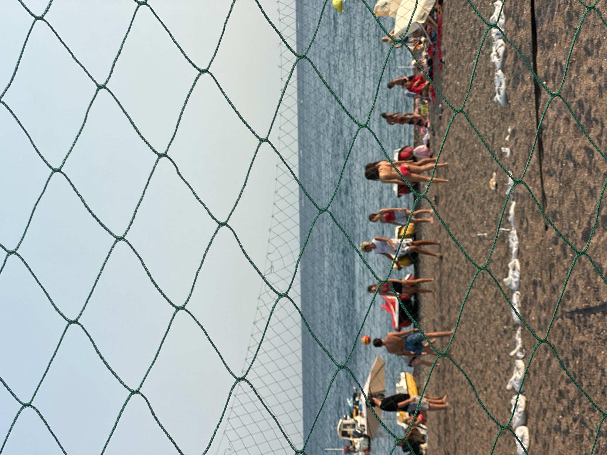 In Boreti, Montenegro, a group of individuals is engaged in a game of beach volleyball on a sandy shoreline. The scene is viewed through a green net, with the net's mesh prominently in the foreground, creating a blurred effect over the background. The ocean stretches out behind the beach, its surface calm under a clear sky. Several small boats are visible near the shore, some pulled up onto the sand. People are scattered on the beach, some participating in the volleyball game, while others appear to be spectating or relaxing. The lighting suggests it is either late afternoon or early morning.