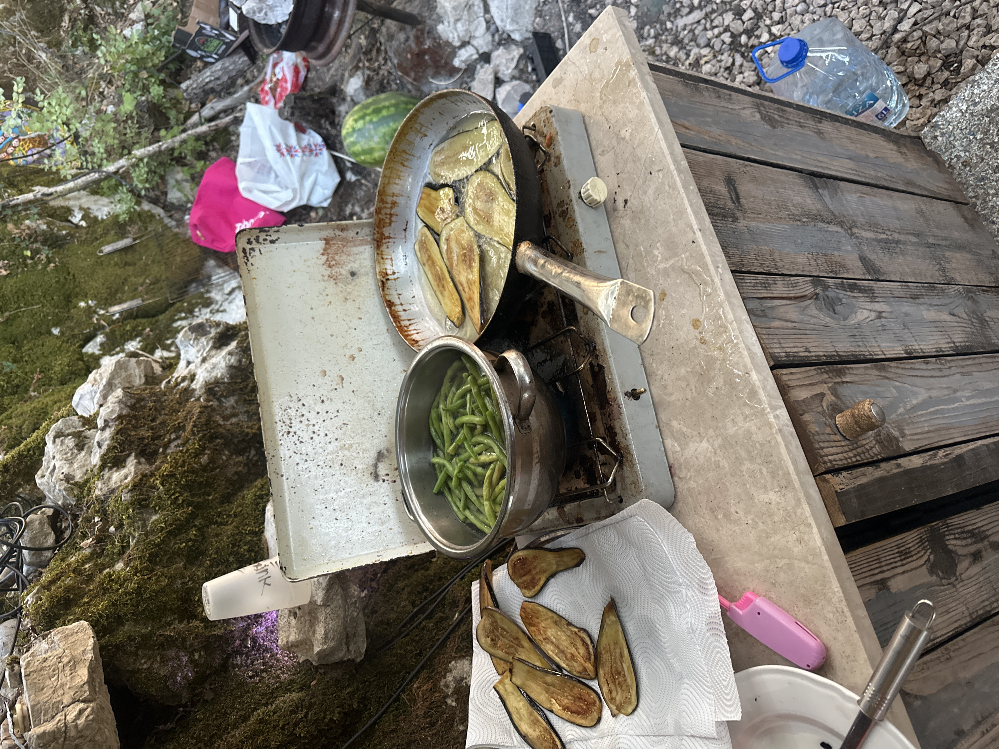 In Bukovik, Montenegro, a portable camping stove is set up on a wooden counter, used for cooking outdoors. Slices of eggplant are being fried in a pan on the right, while green beans are boiling in a pot on the left. Several cooked eggplant slices are laid out on a white paper towel on the left side of the counter. Behind the stove, a watermelon rests on the ground, along with several shopping bags. A large bottle of water is visible on the right side of the wooden counter, near the ground. The scene is set against a backdrop of moss-covered rocks and trees, suggesting a natural, outdoor environment.