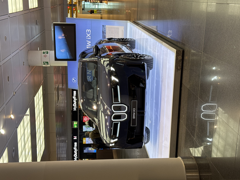 A dark-colored BMW iX3 electric SUV is displayed on a rectangular, illuminated white platform at Munich Airport, Freising, Germany. The vehicle is positioned facing slightly towards the left of the frame, presenting its front fascia, which features an illuminated kidney grille outline and activated headlights. A black panel below the grille prominently states "BMW iX3". Behind the vehicle, a large blue display wall exhibits the BMW logo and "BMW iX3" branding. An overhead monitor, suspended from a white structural pillar, displays dynamic visual content. The dark, polished tiled floor reflects the illuminated elements of the display, including the car's grille lights and the platform's ambient glow. In the background, multiple signs are visible, including "MyDutyFree M" to the left and "Lufthansa Lounge" further to the right, adjacent to an exit sign. Several unidentifiable individuals are observed moving through the terminal; some are near the retail establishments on the left, and one male subject in a yellow shirt is standing further to the right. The environment is an brightly lit indoor airport terminal characterized by a grid of overhead artificial lighting. A large, light-colored structural pillar is partially visible in the left foreground of the image.