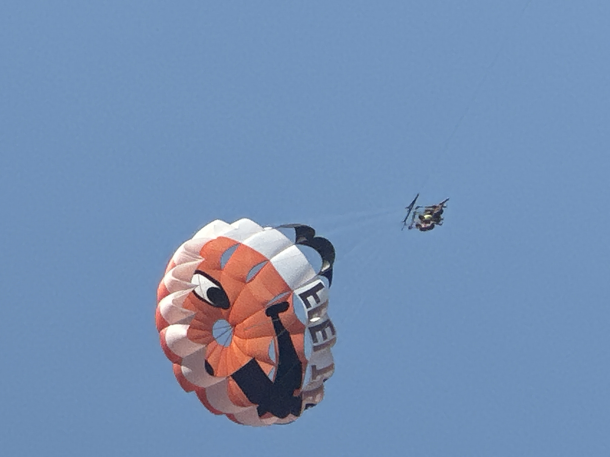 The image shows a parasail in the sky over Boreti, Montenegro. The parasail is orange and white and has a cartoon face on it. Two people are parasailing in the distance. The sky is clear and blue.
