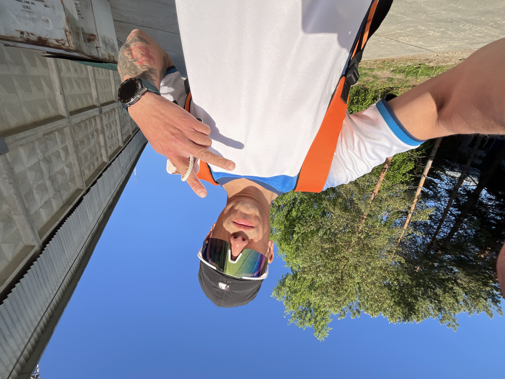 A man is pictured in an outdoor selfie, his face oriented toward the bottom of the frame. He is wearing sunglasses, a baseball cap, and a white t-shirt with orange and black accents. The man makes a "peace" sign with his left hand. On his wrist, he is wearing a watch. A concrete structure is visible to the left, and trees and a clear blue sky are in the background. The image was taken in the city of Chaikovsky, Russia.