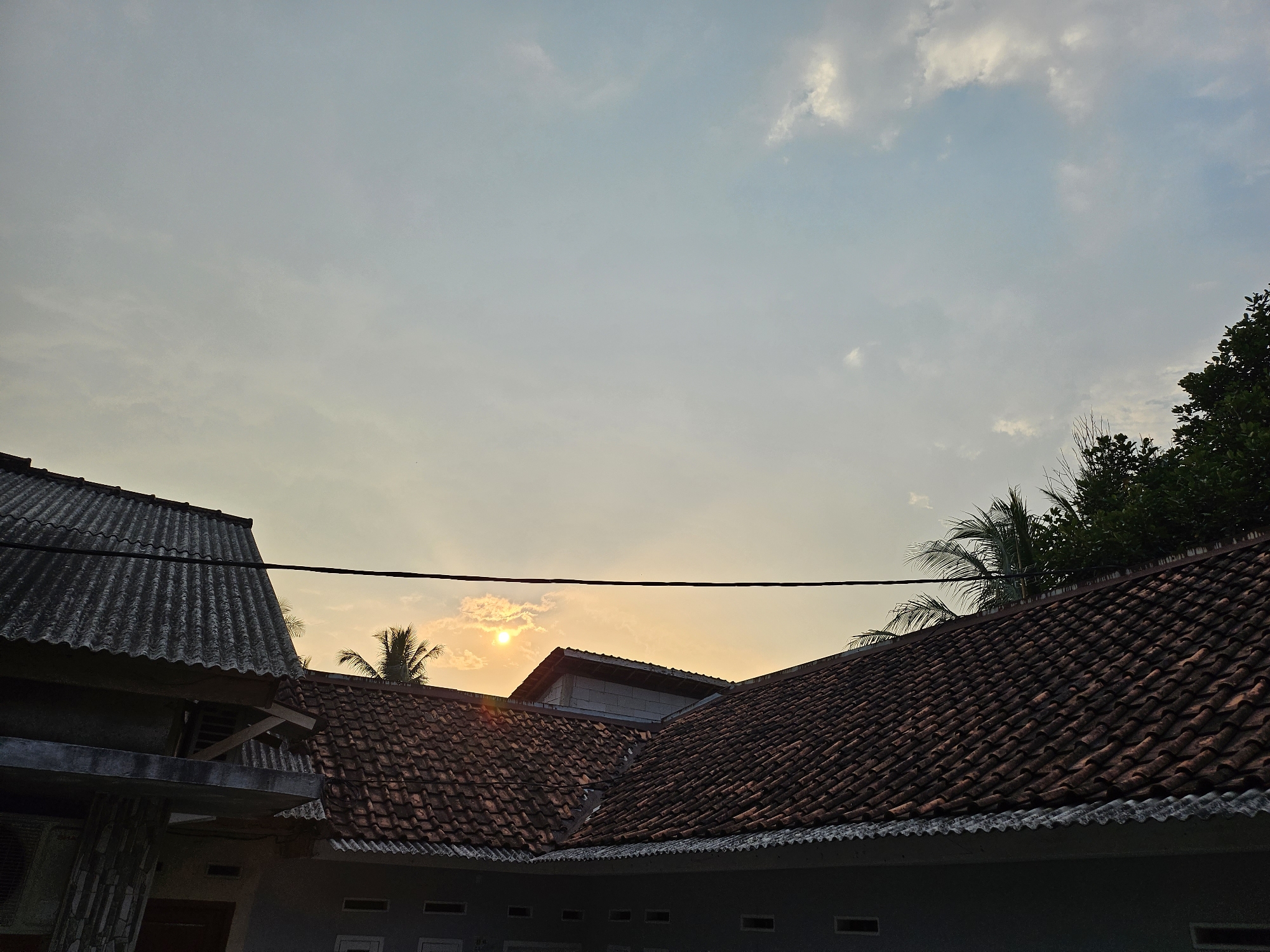 The image shows a view of rooftops and the sky during sunset in Jasinga, Indonesia. In the foreground, there are two distinct roofs: one on the left with a weathered, grey, corrugated material, and another on the right covered with traditional terracotta tiles. A black electrical wire is stretched horizontally across the upper portion of the frame, obscuring a small part of the sky and trees.
The sky is a pale blue with scattered clouds, transitioning to a warm yellow and orange hue near the horizon, indicating the setting sun. The sun itself is visible as a bright, glowing orb partially obscured by a cloud, positioned above and slightly to the left of the center of the image. Several palm trees are visible behind the tiled roof, with their fronds reaching up towards the sky. To the far right, a dense canopy of dark green foliage, likely from larger trees, forms the edge of the scene.
Below the roofs, a portion of a building is visible, characterized by dark grey walls and several rectangular window openings. The overall scene captures a quiet, residential atmosphere during the evening hours. There are no people or animals visible in the frame. The lighting suggests it is late afternoon or early evening.
