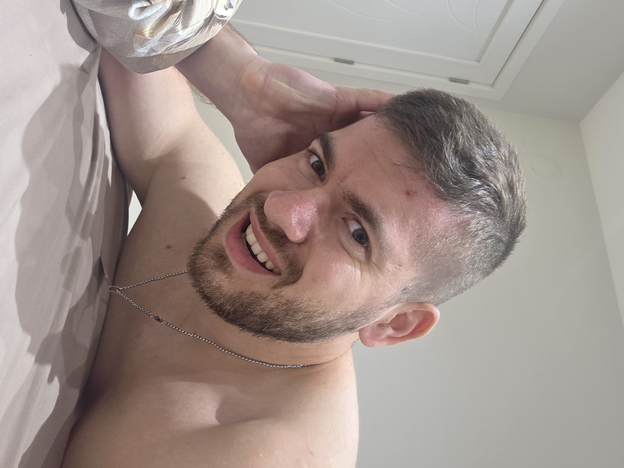 A man lies in bed, smiling at the camera. He has short brown hair, a beard, and is wearing a silver chain. He has a hand on his head. The background is a plain wall and a white ceiling. The location is impossible to determine from the image.