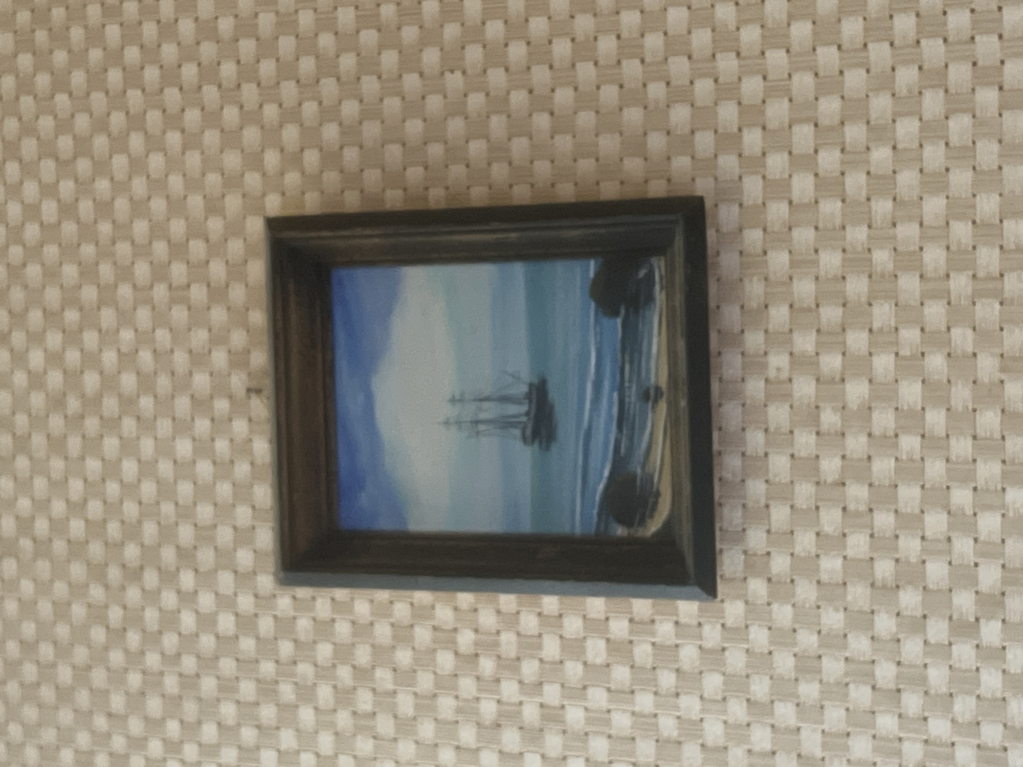 An unframed painting of a sailing ship on the sea is displayed on a light-colored woven textured wall. The painting depicts a dark-hulled ship with three masts sailing on a blue sea under a cloudy sky. The foreground of the painting shows a sandy beach with dark rocks. The painting is hung slightly off-center on the wall. The image does not contain any information about the city of Sudak, Ukraine.