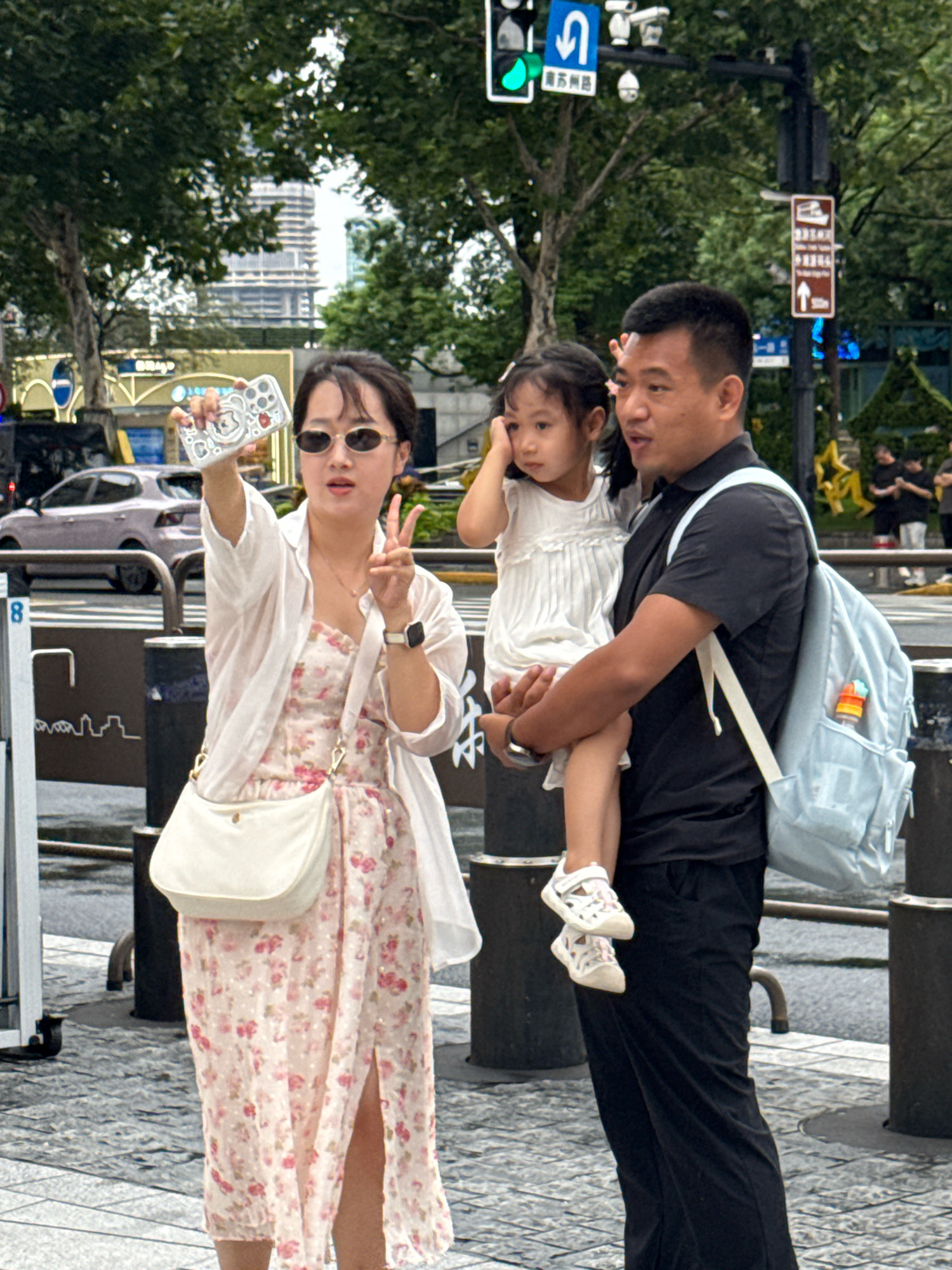 A family of three is taking a selfie on a city street in Shanghai, China. A woman in a floral dress and white cardigan holds a phone up to take the picture, her other hand forming a peace sign. She wears sunglasses and a white crossbody bag. A man holds a young girl, who is wearing a white dress. The man has a light blue backpack slung over one shoulder. The background shows trees, buildings, and street signs. The ground is paved with grey and black square tiles, and a street crossing is visible.