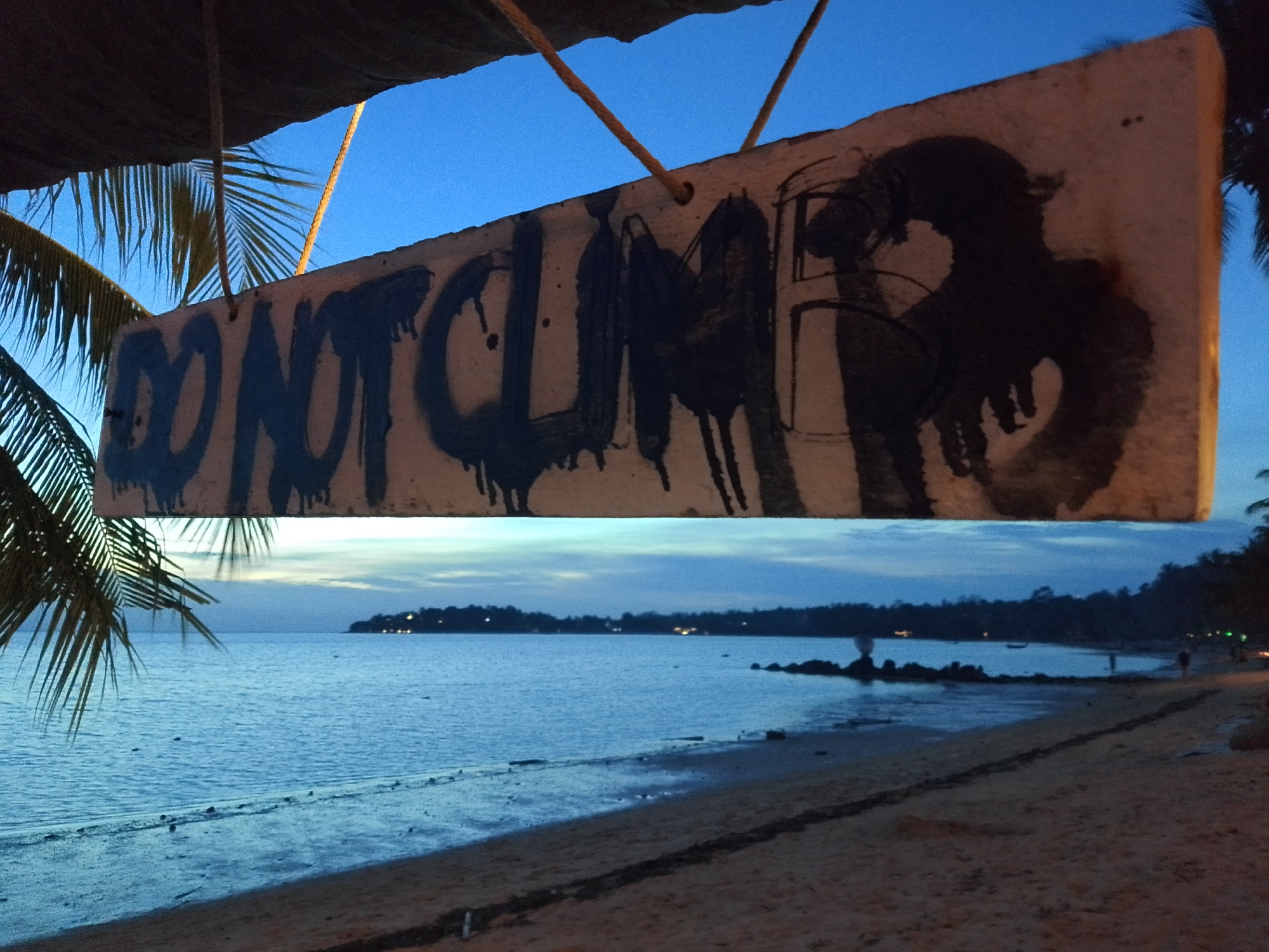An outdoor, medium-shot, eye-level photograph taken at dusk shows a beach scene with a sign in the foreground. The sign, made of a light-colored material with a rough texture, is rectangular and appears to be hanging from ropes. It is marked with large, dark spray-painted letters that read "DO NOT CLIMB." To the right of the text, a silhouette of what appears to be an animal or a mythological creature is spray-painted in black. The sign is positioned against a clear blue twilight sky, with palm fronds visible on the left side, partially obscuring the sign and the sky.
Below the sign, a wide expanse of ocean stretches to the horizon. The water is calm and reflects the dim light of dusk. A sandy beach runs along the edge of the water, with some debris and vegetation scattered on it. In the distance, a dark, tree-lined coastline is visible, with a few small lights suggesting human habitation. The overall atmosphere is serene and somewhat moody, characteristic of the transition from day to night. The specific location is described as ตำบลเกาะพะงัน, Thailand, suggesting a tropical or subtropical coastal environment. There are no people clearly visible in the foreground or mid-ground, though a few figures might be present on the distant shore. The image captures a moment of quiet beauty, with the stark warning on the sign adding a touch of intrigue to the peaceful landscape.