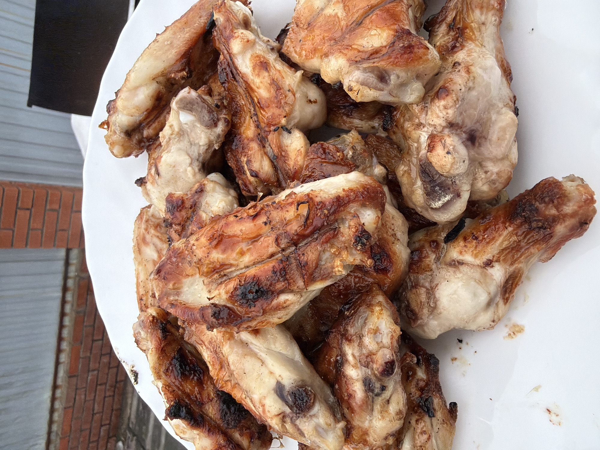 The scene depicts a white, round platter containing approximately 20-30 cooked chicken pieces, primarily wings and wing segments, arranged in a pile. The chicken is golden-brown with areas of dark charring, indicative of grilling or roasting. Grease and small food particles are visible on the white platter. The setting is outdoors in Chaikovsky, Russia. In the background, a red brick wall is partially visible on the left, transitioning to a light grey corrugated metal fence or wall in the center. A dark, rectangular object, possibly a grill, is partially visible on the far right behind the metal barrier. The ground beneath the brick wall appears to be a dark, possibly paved or earthen surface. No individuals, actions, or specific interactions are visible.