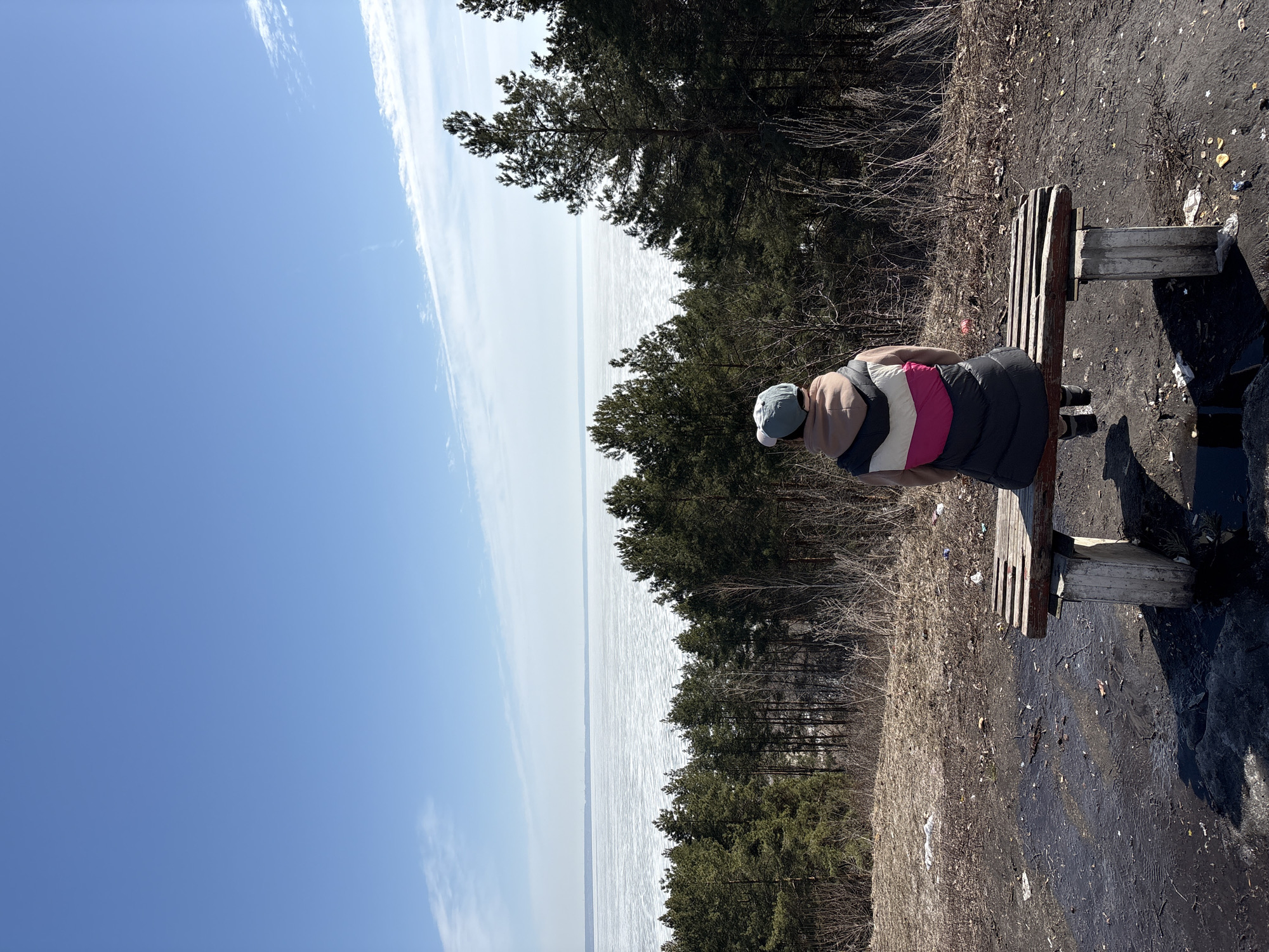 A person is sitting on a wooden bench facing a body of water in a location with many trees and a mostly clear sky. The water appears to be frozen. The ground around the bench is littered with trash. This scene is in the city of Ulyanovsk, Russia.
