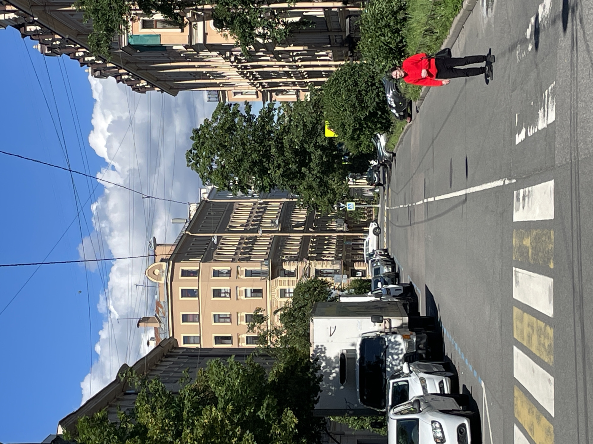 The image depicts a street scene in Saint Petersburg, Russia, on a sunny day with scattered clouds. The street is lined with multi-story buildings featuring neoclassical architecture. There are several cars parked along the street, including a white truck, several white passenger cars, and a row of darker colored cars. A pedestrian wearing a red sweatshirt and black pants is standing in the middle of the street near a crosswalk. There are overhead power lines, indicating it may be a streetcar route. Lush green trees and bushes are present along the sidewalks and between buildings, adding a touch of nature to the urban environment. The overall impression is of a typical city street during daylight hours.