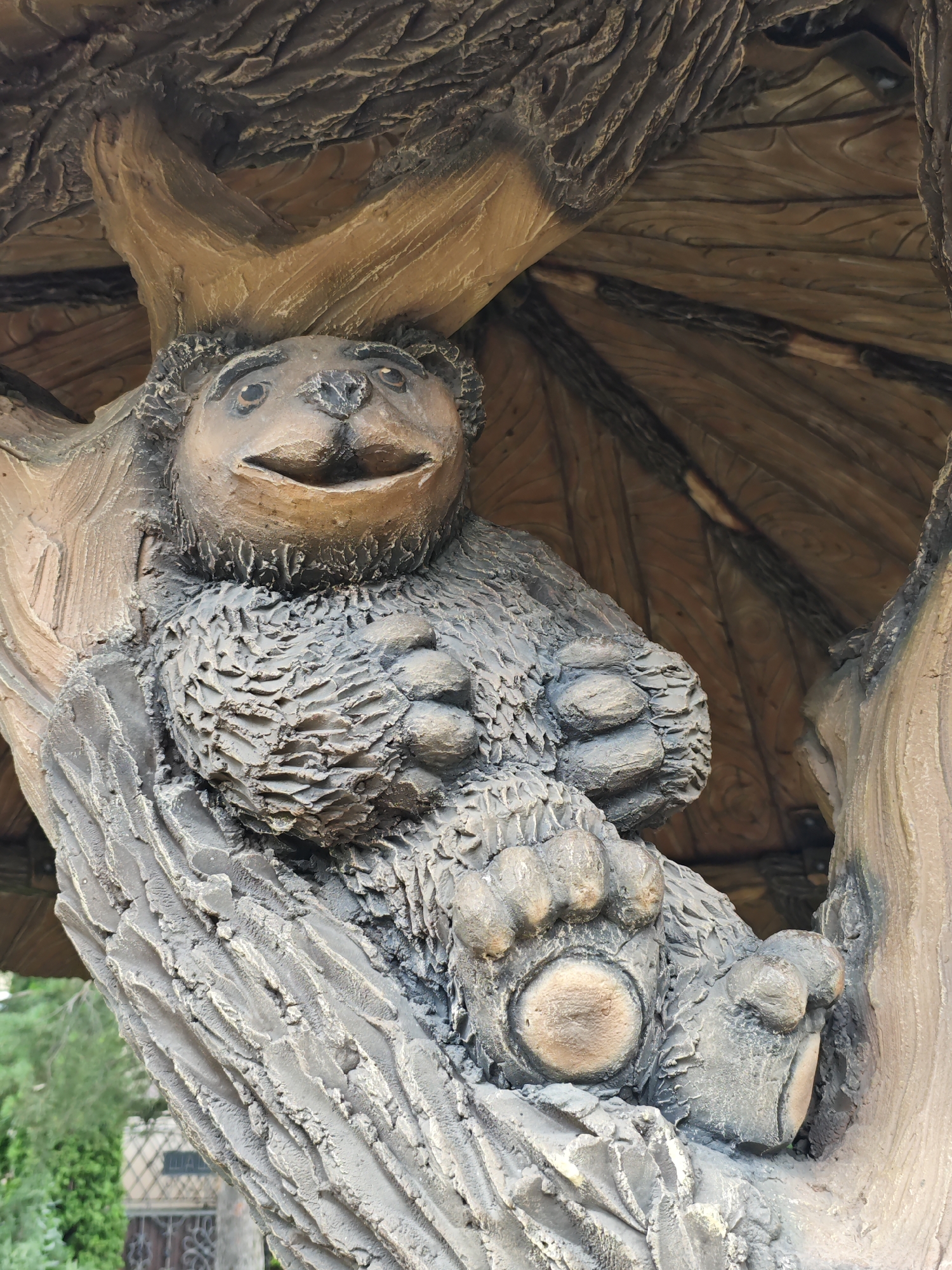 A statue of a bear is carved into a tree trunk, appearing to be seated with its paws resting on the wood. The bear is depicted with a wide smile and textured fur. The background shows a wooden ceiling structure with a rough, bark-like texture. The environment appears to be outdoors, with hints of greenery and a fence visible in the distance. This sculpture is located in Cherkessk, Russia, likely within a park or public area, as suggested by the surrounding vegetation and architectural elements.