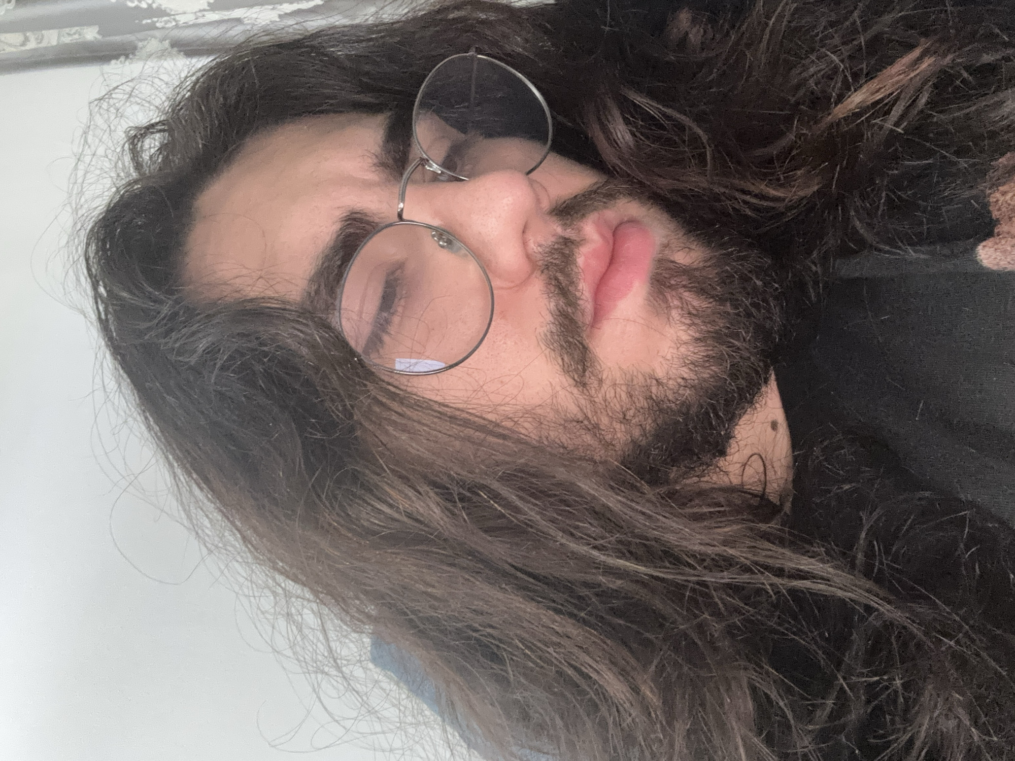 A close-up, overhead shot shows a person with long, dark, wavy hair and a beard, wearing round, silver-rimmed glasses. The person's face is partially obscured by their hair and glasses. Their eyes are closed, and their lips are slightly parted. The person is wearing a black shirt. The background is out of focus and appears to be a plain wall and a curtain. The lighting is soft, suggesting an indoor setting, possibly a bedroom or living room. The image is a selfie, indicated by the angle and the close-up framing of the face. There are no contextual clues to suggest a specific city or location.