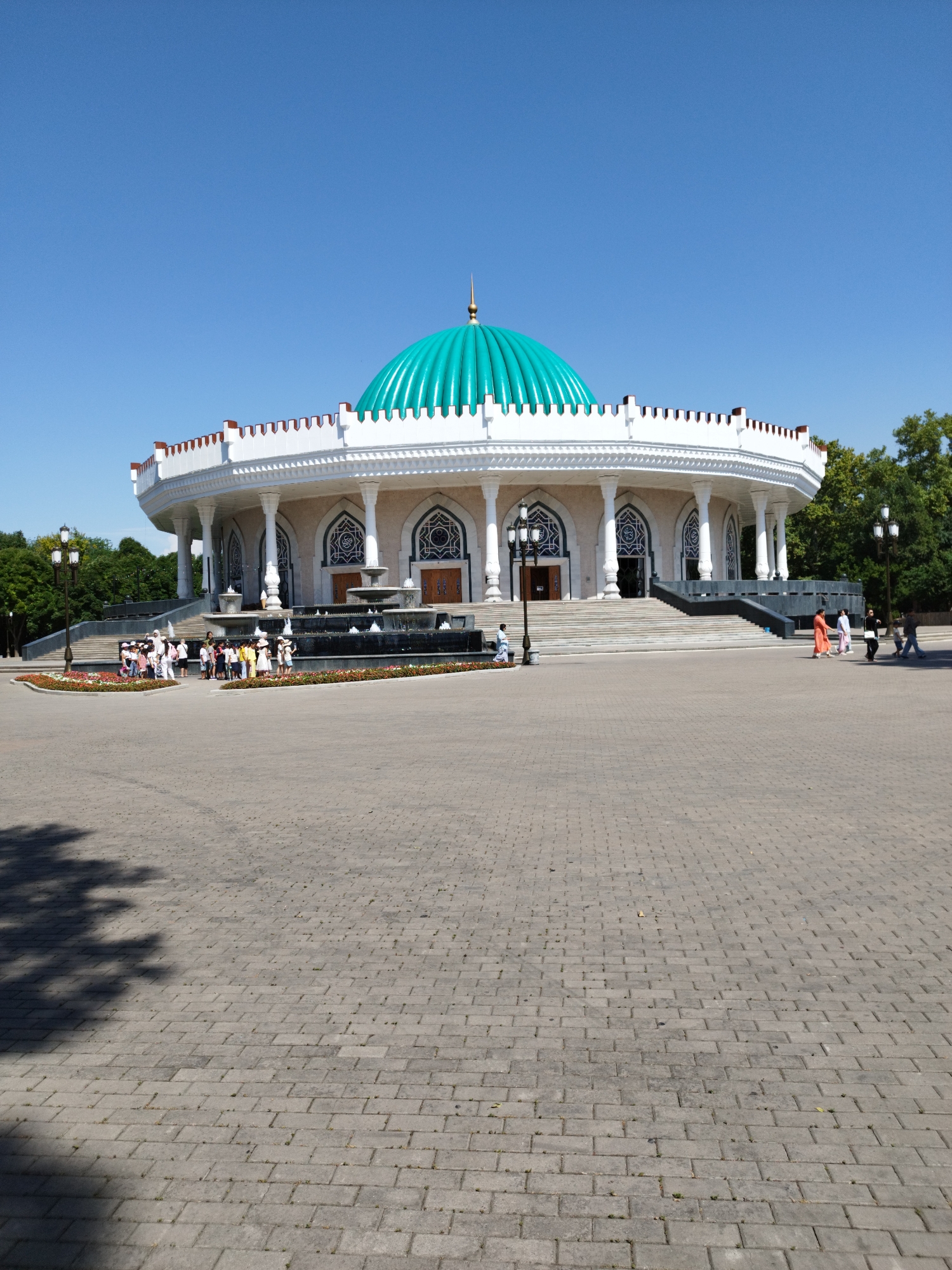 The image shows a wide-angle view of a grand, circular building with a prominent teal-colored dome, topped with a golden spire. The building is white with ornate columns and decorative arched windows. A series of fountains are visible in front of the building. Several people are gathered in front of the building, some standing in a group and others walking across the paved plaza. The plaza is made of grey interlocking pavers. Trees and greenery are visible behind the building and to the sides. The sky above is a clear, bright blue.