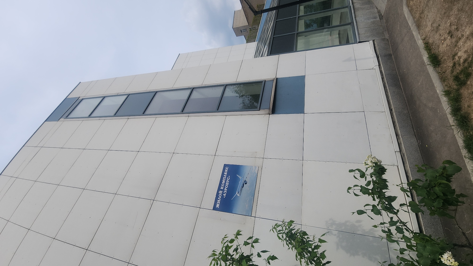 A low-angle shot shows the exterior of a building in Moscow, Russia. The building has a white tiled facade with blue accents around the windows. A sign is affixed to the wall, reading "ЖИЛОЙ КОМПЛЕКС АЭРОБУС" (ZHILOY KOMPLEKS AEROBUS) with an image of an airplane on a blue background. Plants grow near the base of the building, and a concrete walkway runs along the side. Another building with windows is visible in the background. The sky is visible at the top of the image.