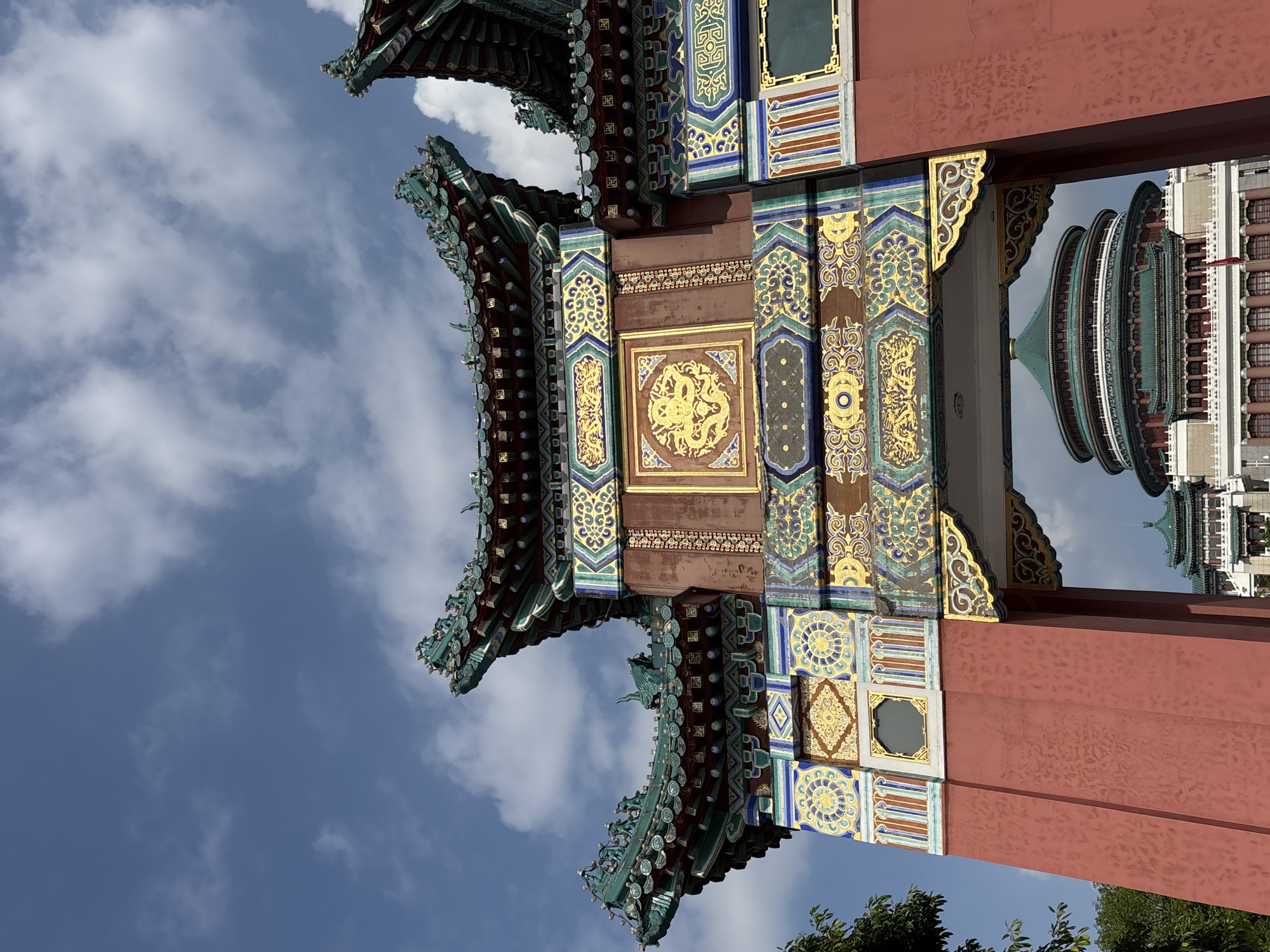 This is a close-up, low-angle shot of the Hongyadong complex in Yuzhong District, China, focusing on an ornate, traditional Chinese archway. The archway is painted a deep red and is elaborately decorated with intricate patterns in gold, green, blue, and yellow. A prominent gold dragon motif is centered on the archway's facade. The roof of the archway features traditional Chinese architectural elements, with curved eaves and green glazed tiles, adorned with small figurines. Behind the archway, through its opening, a large, multi-tiered, circular building with a green tiled roof and white walls is visible, also featuring traditional Chinese design. The sky above is a clear blue with scattered white clouds. The overall scene depicts elements of traditional Chinese architecture within a modern urban setting.