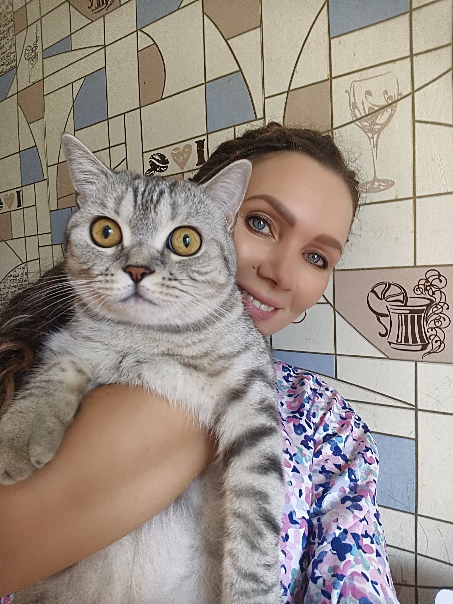 A young woman with braided hair and blue eyes smiles at the camera while holding a gray tabby cat. The woman is wearing a colorful floral print shirt. The cat has large yellow eyes and is looking directly at the camera. The background is a tiled wall with a geometric pattern and images of a wine glass and a cocktail. The scene appears to be indoors, possibly in a home.