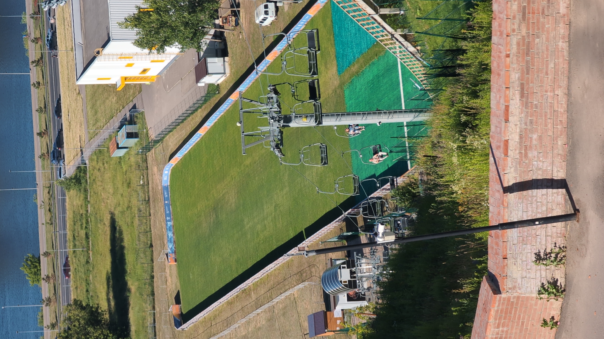 This is an aerial photograph of a ski slope with a ski lift in Nizhny Novgorod, Russia. The ski slope is covered in green grass, suggesting it is either summer or a man-made ski slope designed for year-round use. There are several ski lift chairs suspended on a cable, with a few people visible riding in some of them. The ski lift extends upwards and to the right of the frame.
In the background, there is a building with a white and yellow facade, and to the left of that, a body of water, likely a river or canal, is visible. A road with several cars is running parallel to the water. Further to the right of the ski slope, a brick wall runs along the edge of the frame, casting a shadow onto a paved path. The scene appears to be bathed in sunlight, indicating a clear day, likely midday or early afternoon. The presence of a ski lift and a prepared slope implies that this is a recreational area, possibly a sports complex. There is no visible text in the image.