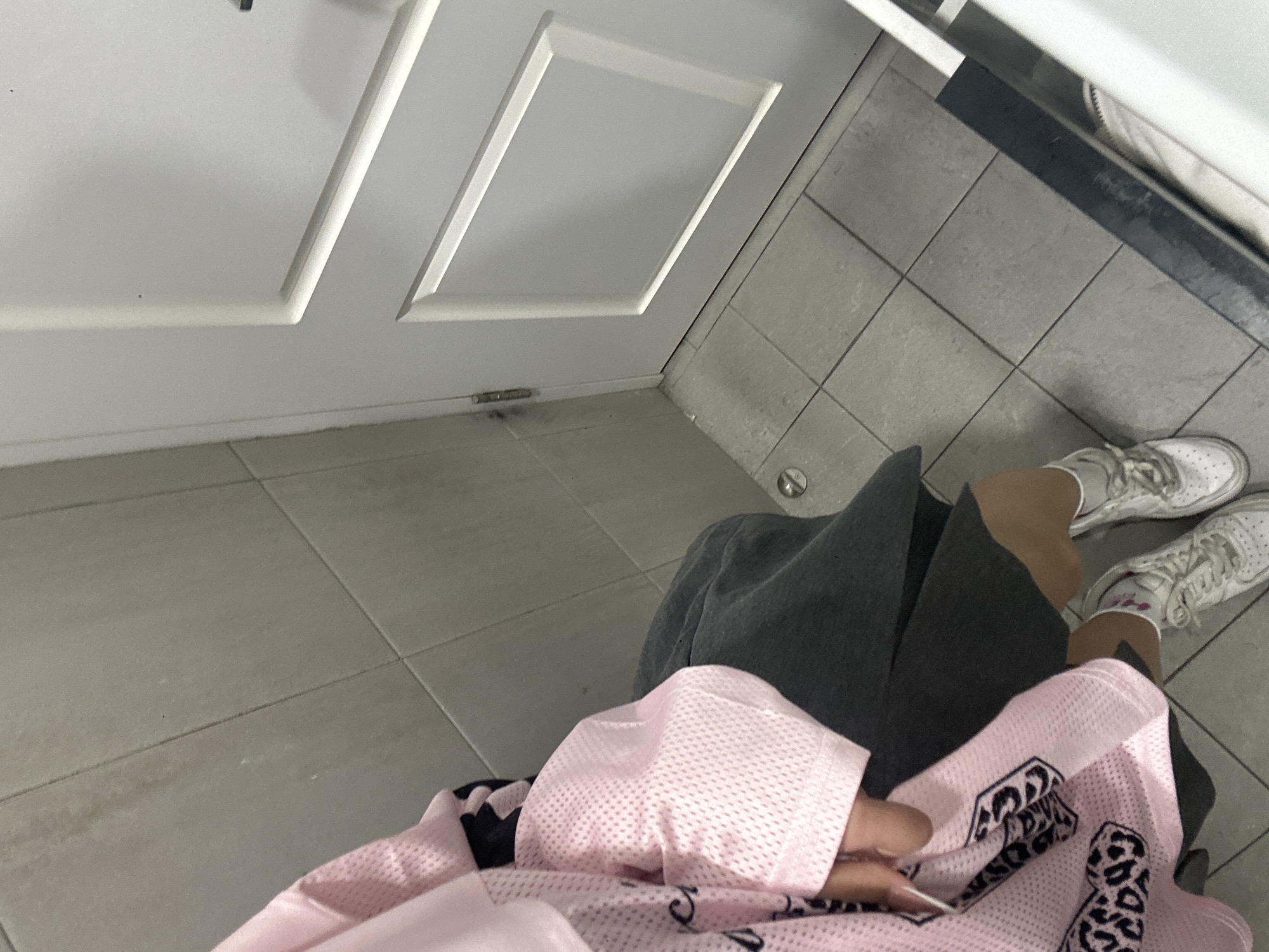The image is captured from a high-angle, downward perspective, showing a person's lower body, their clothing, and the immediate indoor environment.
The setting appears to be an interior space, possibly a hallway or room, featuring light gray square floor tiles with darker grout lines. Some areas of the tiles exhibit minor discoloration or scuff marks, suggesting normal wear and tear. To the left, there is a white wall or door with raised rectangular panels, indicating a traditional design. A dark hinge or mark is visible near the bottom of this white surface, confirming it might be a door. Along the right edge of the frame, a darker, possibly black or dark gray, horizontal surface runs parallel to the tiles, which could be the base of a wall, a counter, or a piece of furniture.
The person in the image is seen wearing dark gray shorts or loose-fitting pants that extend to about mid-thigh or knee-length. Their legs, with a light to medium skin tone, are visible below the shorts. On their feet, they wear white athletic shoes with laces and white socks. A faint pink pattern or logo is visible on one of the white socks.
The person is holding or wearing a pink, mesh-like fabric garment, possibly a jersey or a light jacket. The fabric has a distinct porous texture, typical of athletic wear. A prominent black design is visible on the pink fabric, featuring stylized text or a logo. While difficult to decipher completely due to the angle and partial view, parts of the design include rounded shapes, possibly letters like "C," and what appears to be a leopard print or spotted pattern integrated into some of the elements. It's a bold and distinctive graphical design. A hand with long, possibly manicured, nails is visible holding the pink garment.
The lighting in the image appears to be artificial indoor lighting. No specific time of day, weather, or discernible emotions are apparent.