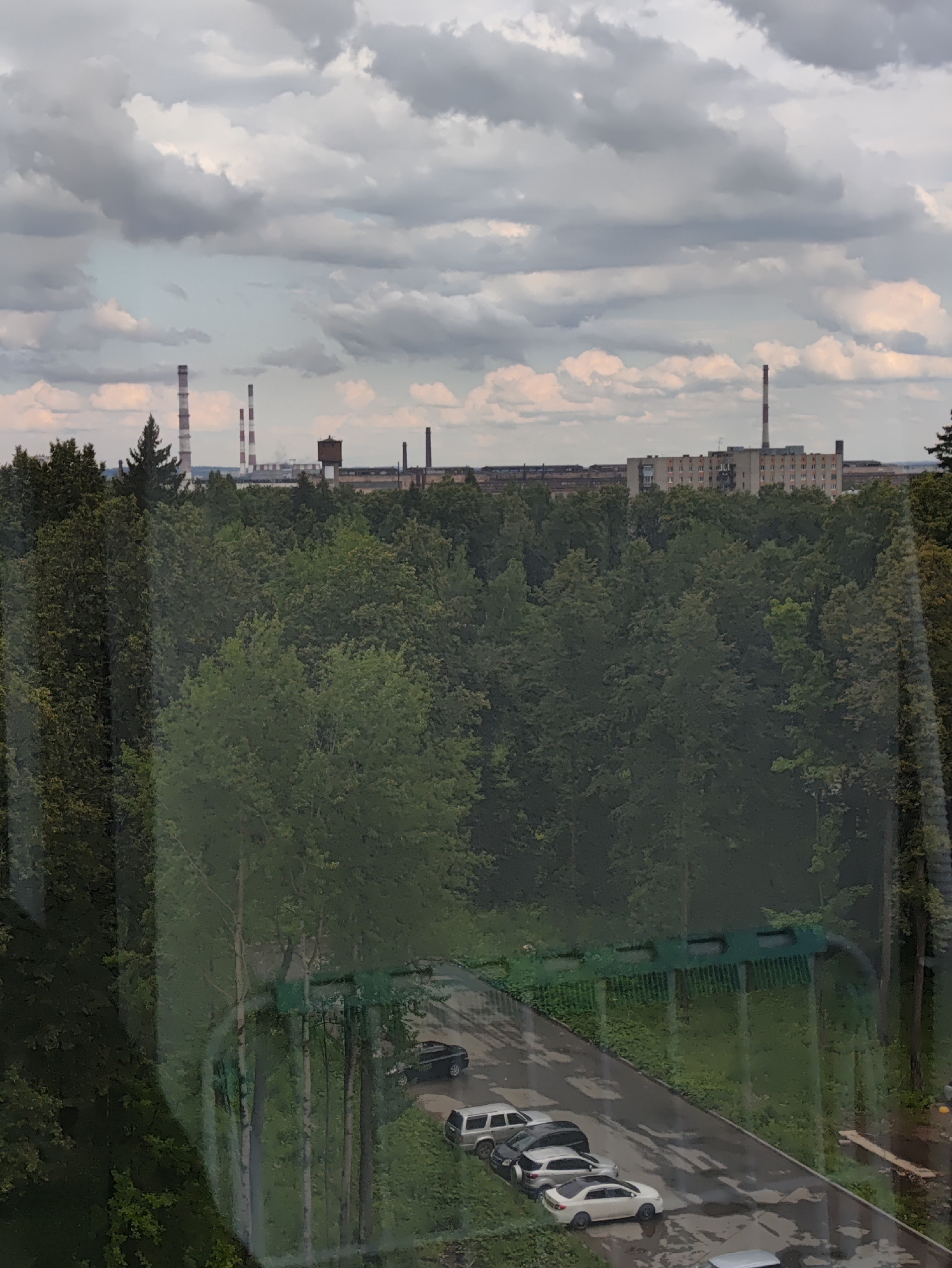 The image shows a view of an industrial area and residential buildings in Izhevsk, Russia. In the background, several tall industrial smokestacks, likely part of a power plant or factory, are visible against a cloudy sky. In the middle ground, a dense forest of green trees dominates the landscape. In the foreground, a parking lot with several cars, including a white sedan, a dark sedan, and a silver SUV, is visible. The ground appears wet, suggesting recent rainfall. The scene is likely a residential area in Izhevsk, overlooking an industrial zone and a forested area.