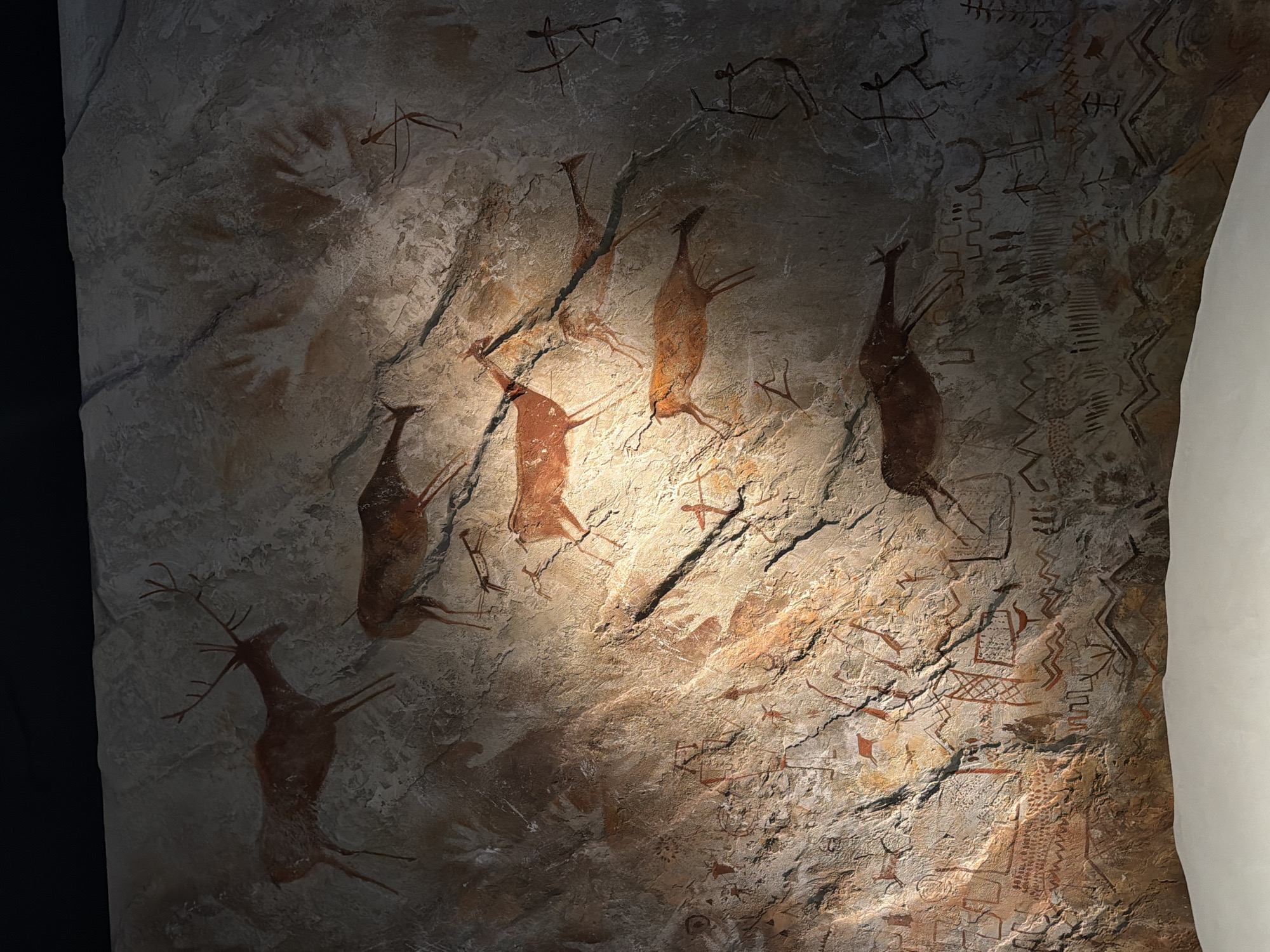 The image shows a rock wall with cave paintings. The paintings depict animals, including deer, and abstract symbols. Handprints are also visible. The style and subject matter suggest these are prehistoric artworks. The location of the image is not discernible.