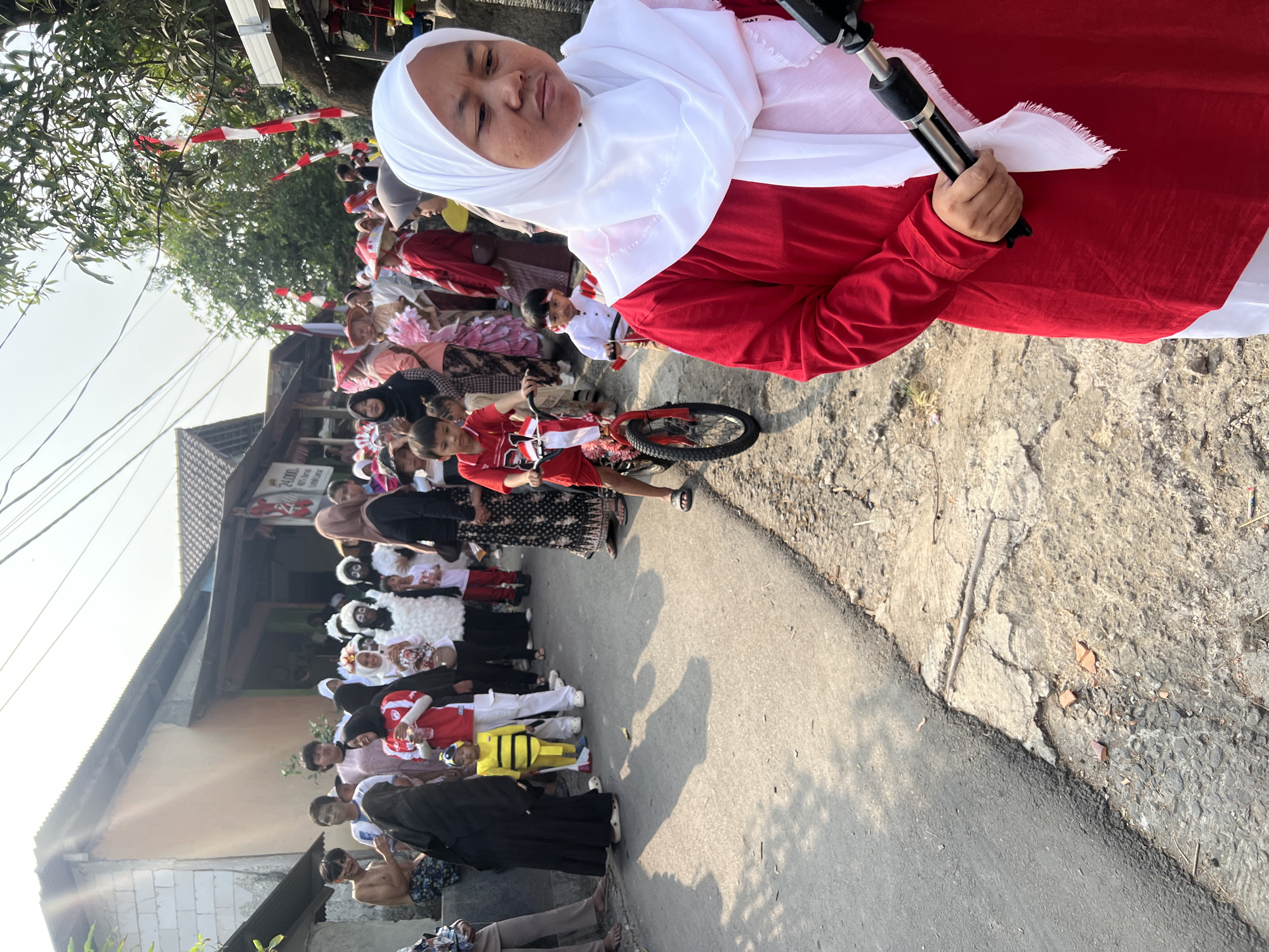A group of people, including children, are gathered on a street in Klapanunggal, Indonesia. A young boy in a red shirt and white shorts rides a small bicycle adorned with Indonesian flags. To his right, another young boy in a white shirt and brown shorts is also on a bicycle. In the foreground, a woman wearing a red shirt and a white hijab is holding a camera on a monopod. Several other individuals, dressed in various traditional and costume attire, are visible in the background, some wearing white fluffy costumes and others in colorful outfits. The scene appears to be a local celebration or parade, with Indonesian flags decorating the street and buildings. The environment includes paved road, cracked concrete sidewalks, and houses with corrugated metal roofs. The sky is bright and sunny.