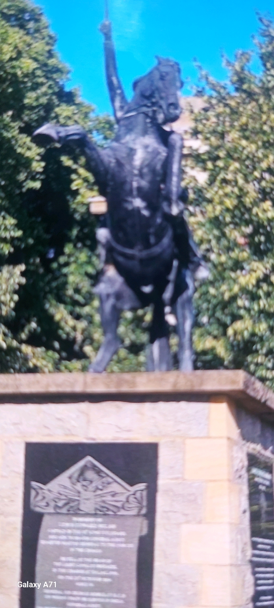 This is a low-resolution, slightly blurry image of a monument in Moscow, Russia. The monument features a dark bronze equestrian statue of a figure, possibly a military leader, on horseback. The rider's arm is raised, holding what appears to be a sword or lance. The horse is depicted in mid-stride, with its front legs elevated. The statue is situated on a stone pedestal, which has a plaque on its front. The plaque contains text and an engraved decorative element at the top, possibly a crest or emblem. Behind the monument, lush green trees are visible against a clear blue sky. The lighting suggests it is daytime. The "Galaxy A71" watermark in the bottom left corner indicates the image was captured with a Samsung Galaxy A71 phone.