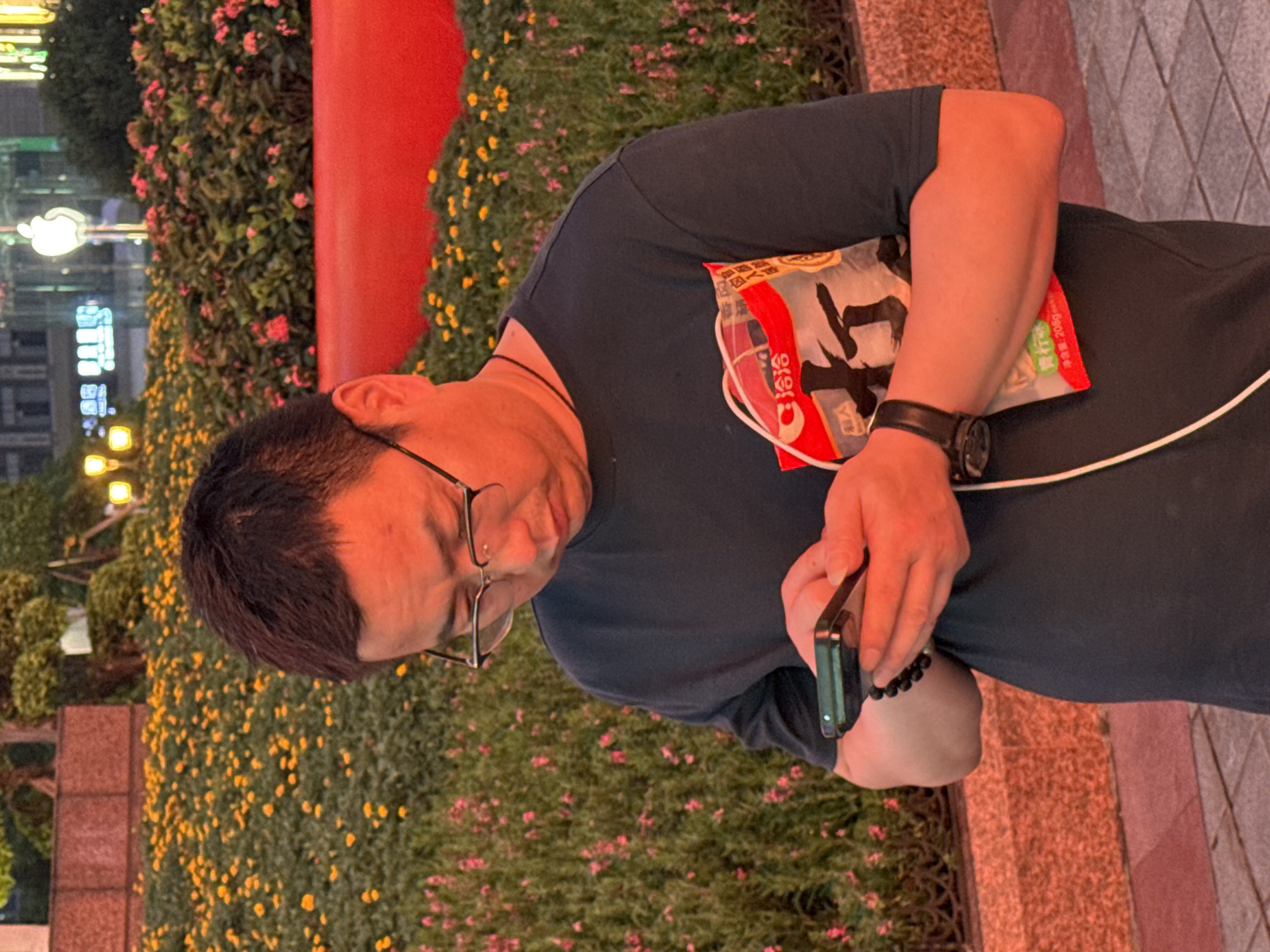 A medium shot captures a man with dark hair and glasses, wearing a dark t-shirt and a black watch, standing outdoors and looking down at his smartphone. He holds a red and white bagged item with Chinese characters in his left arm. The background features a lush green hedge with yellow and pink flowers, and a vibrant red structure. The scene is illuminated by soft lighting, suggesting it might be evening or a shaded area, with building lights visible in the distance. The man's posture and focus on his phone indicate he is engaged in a digital activity.