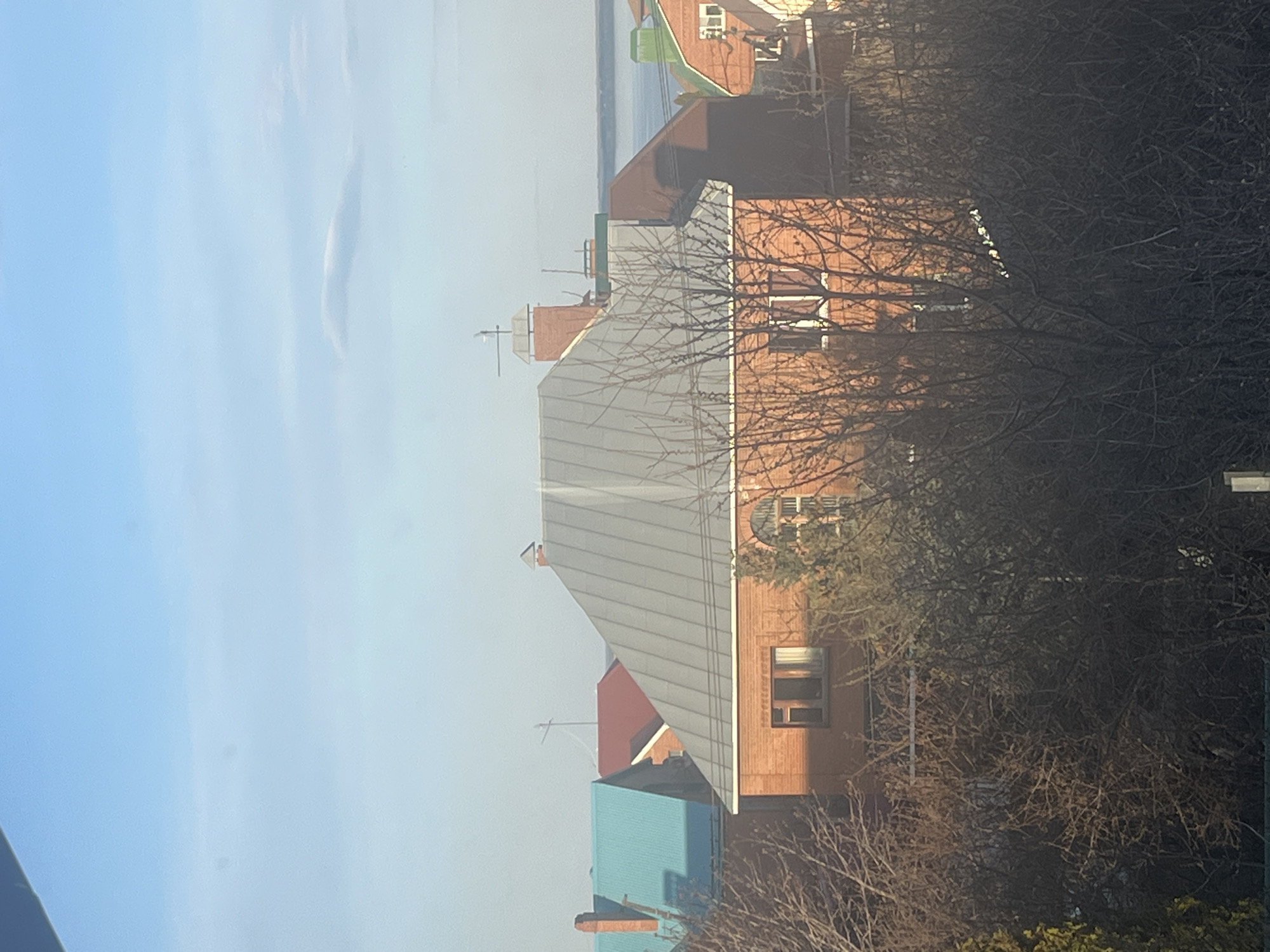 The image depicts a residential area in Ulyanovsk, Russia, possibly photographed from a window overlooking a neighborhood. Several multi-story brick buildings with grey metal roofs are visible. Some buildings have distinctive architectural features, such as rounded windows. Bare trees are prominent in the foreground, partially obscuring some of the buildings. The sky is light blue with a faint, elongated cloud formation. In the distance, a body of water (likely the Volga River) is visible. The overall scene is bathed in sunlight, casting shadows on the buildings and creating a sense of a clear, possibly cold day.